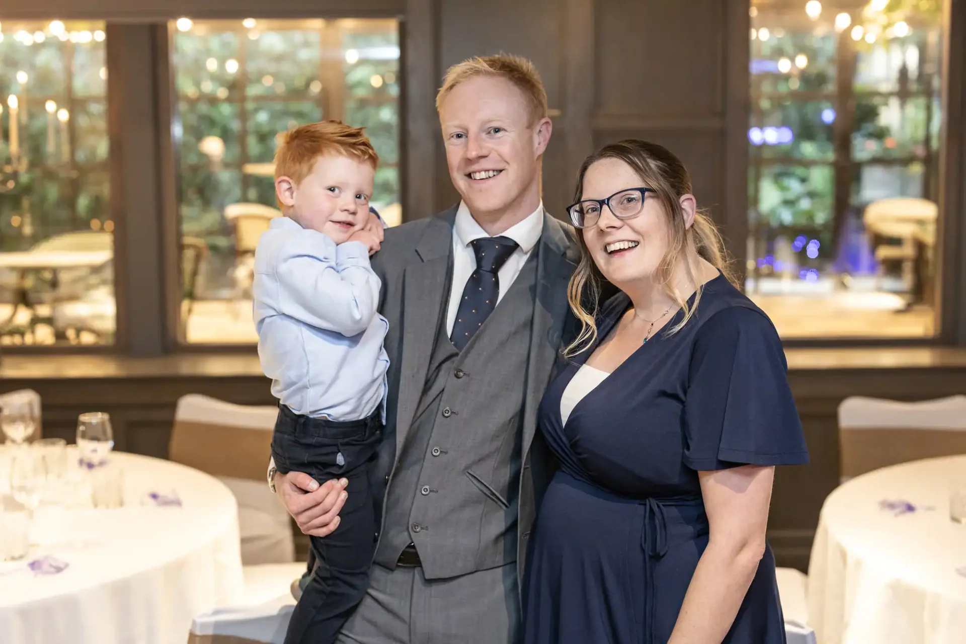 A man in a suit holds a young boy while standing next to a woman in a blue dress. They are smiling, with banquet tables and soft lights in the background.