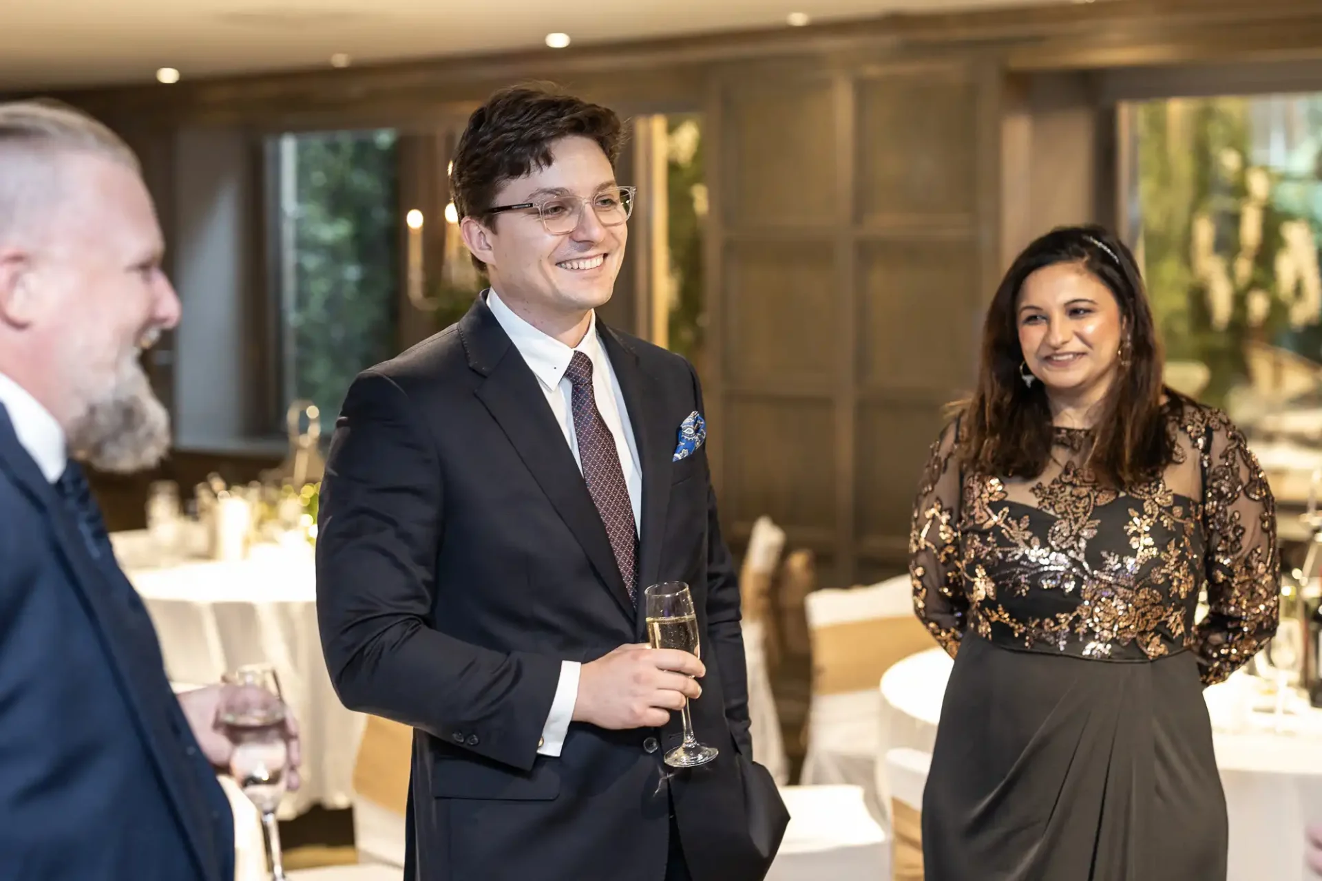 Three people in formal attire stand indoors, two holding drinks, engaged in conversation and smiling.