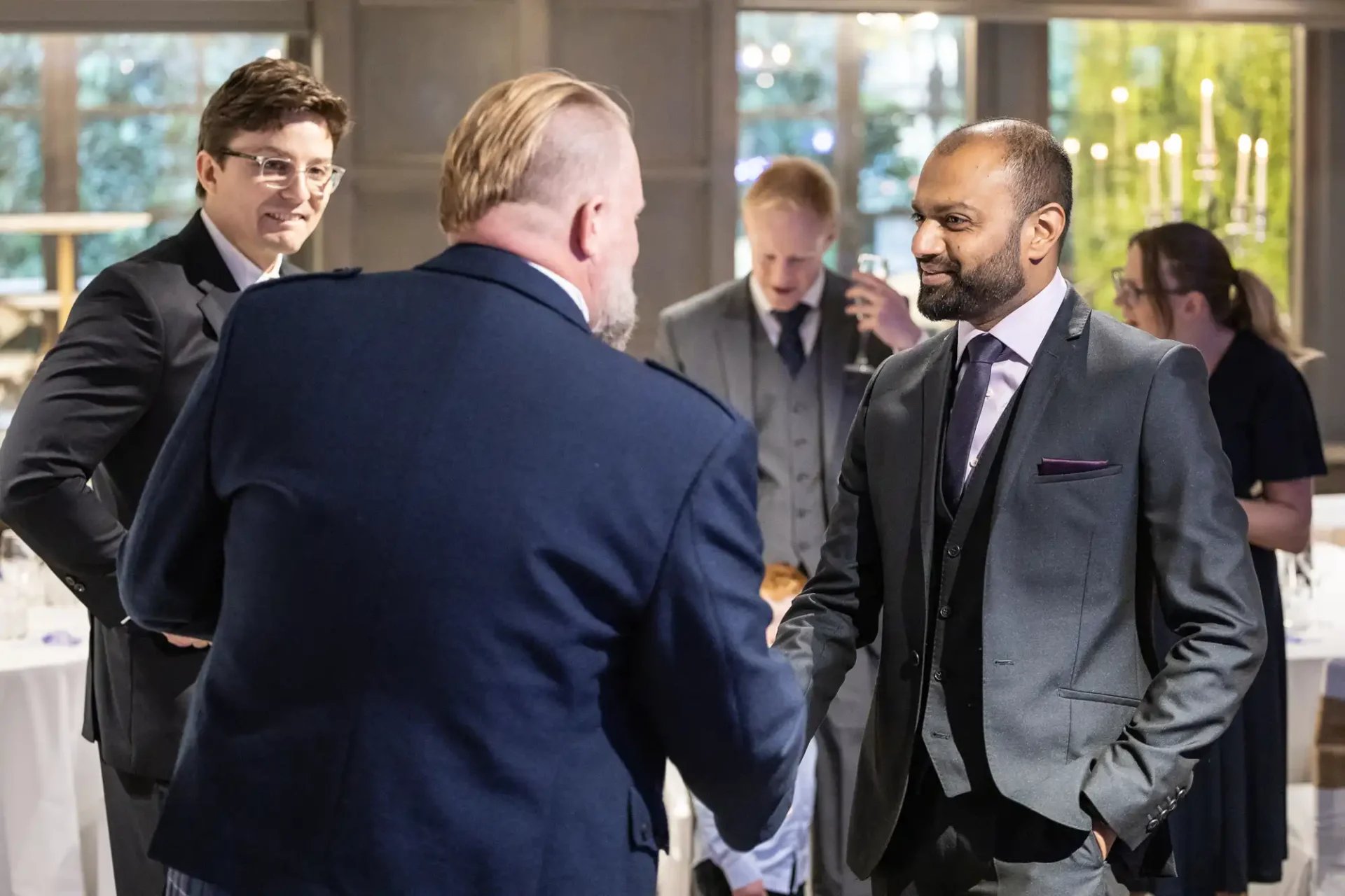 A group of people in formal attire are engaged in conversation and networking at an indoor event, with one person shaking hands.