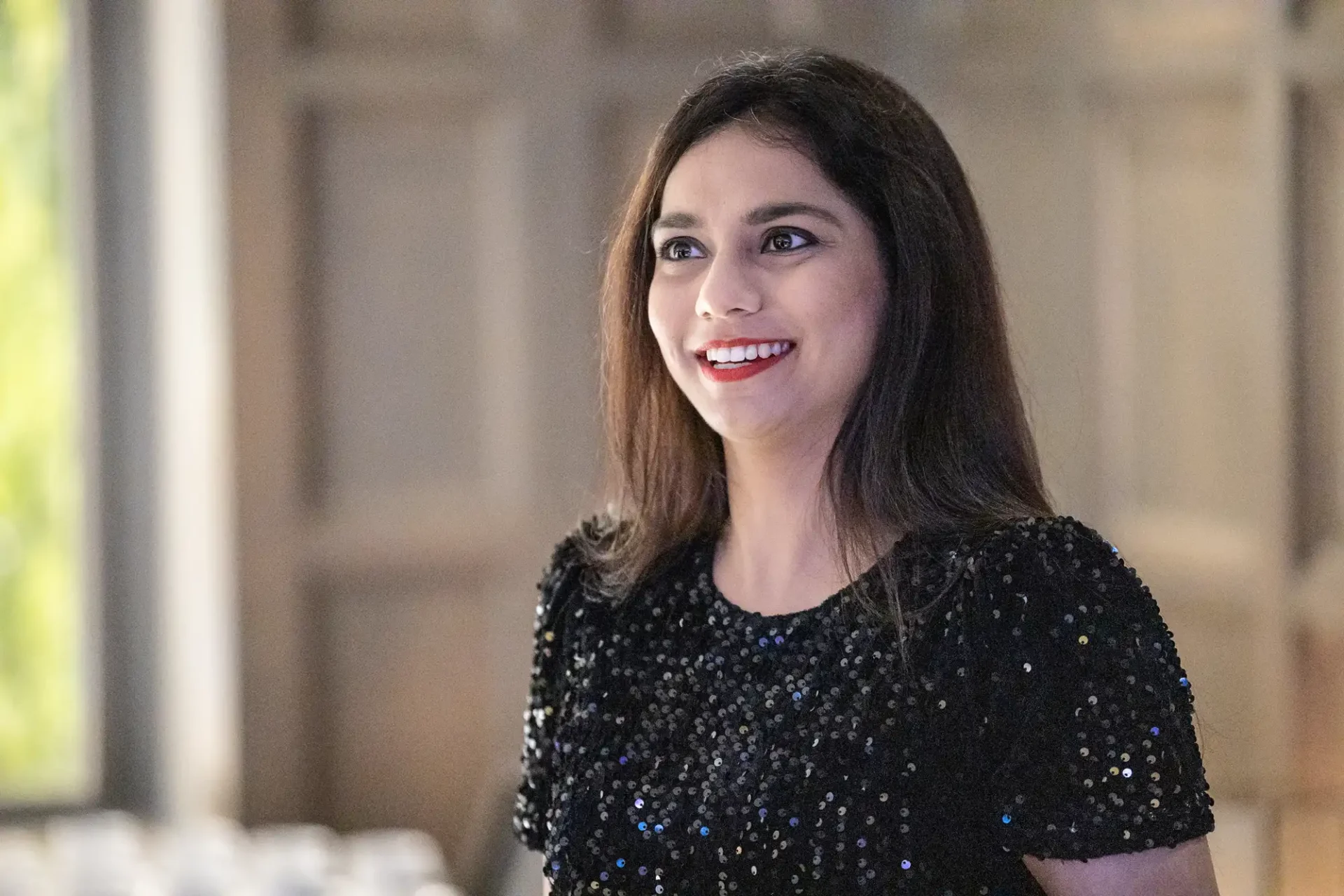 A woman with long dark hair smiles while wearing a sequined black top, standing indoors with a blurred background.