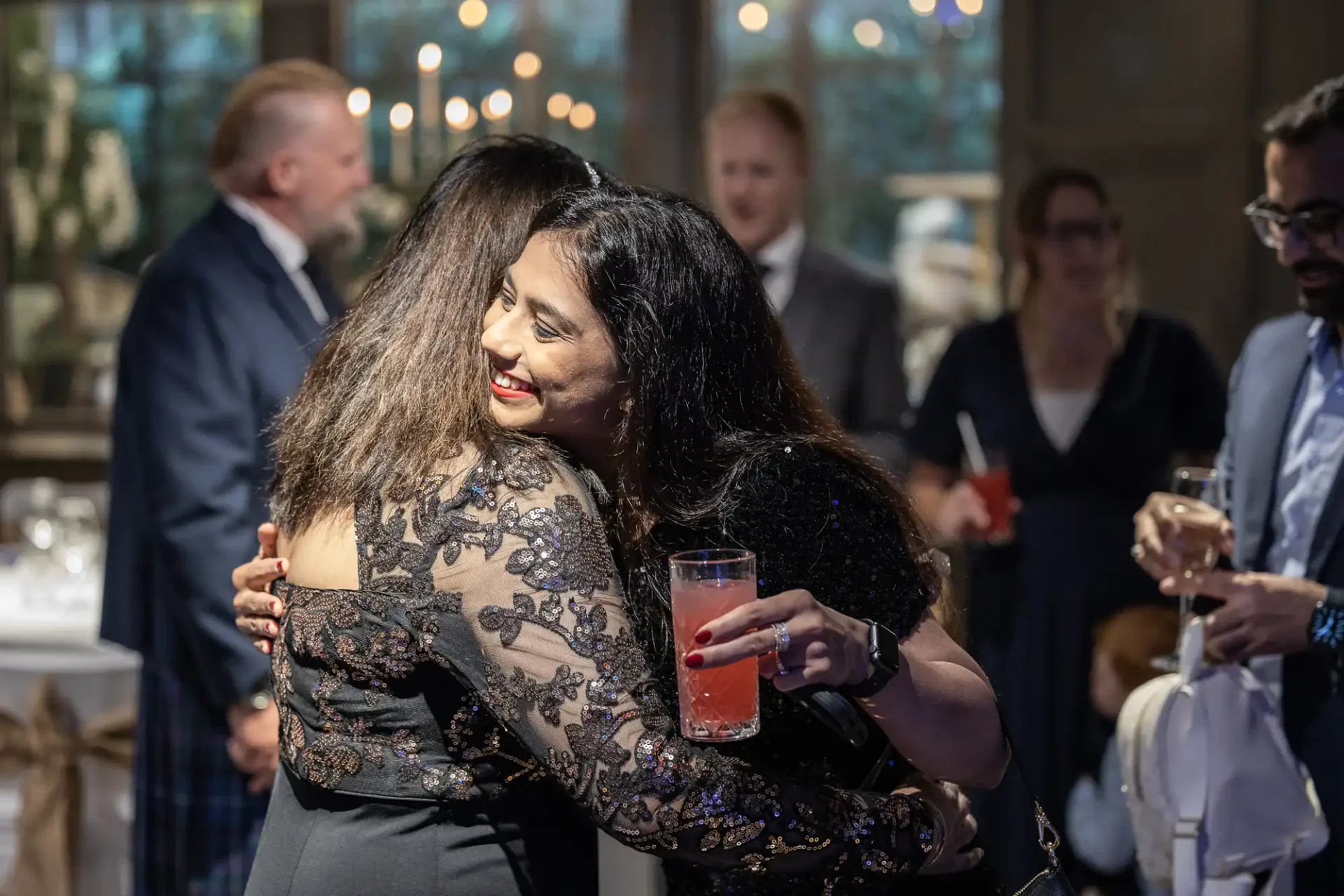 Two women in formal attire share a hug at a social gathering. One holds a pink cocktail. People in the background are engaged in conversation.