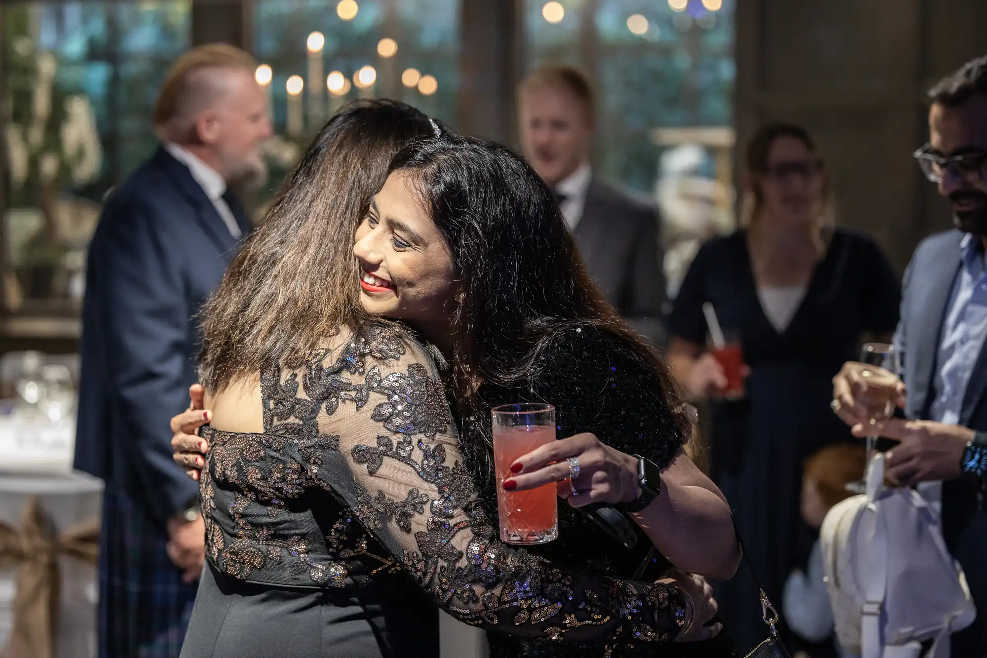 Two women in formal attire share a hug at a social gathering. One holds a pink cocktail. People in the background are engaged in conversation.