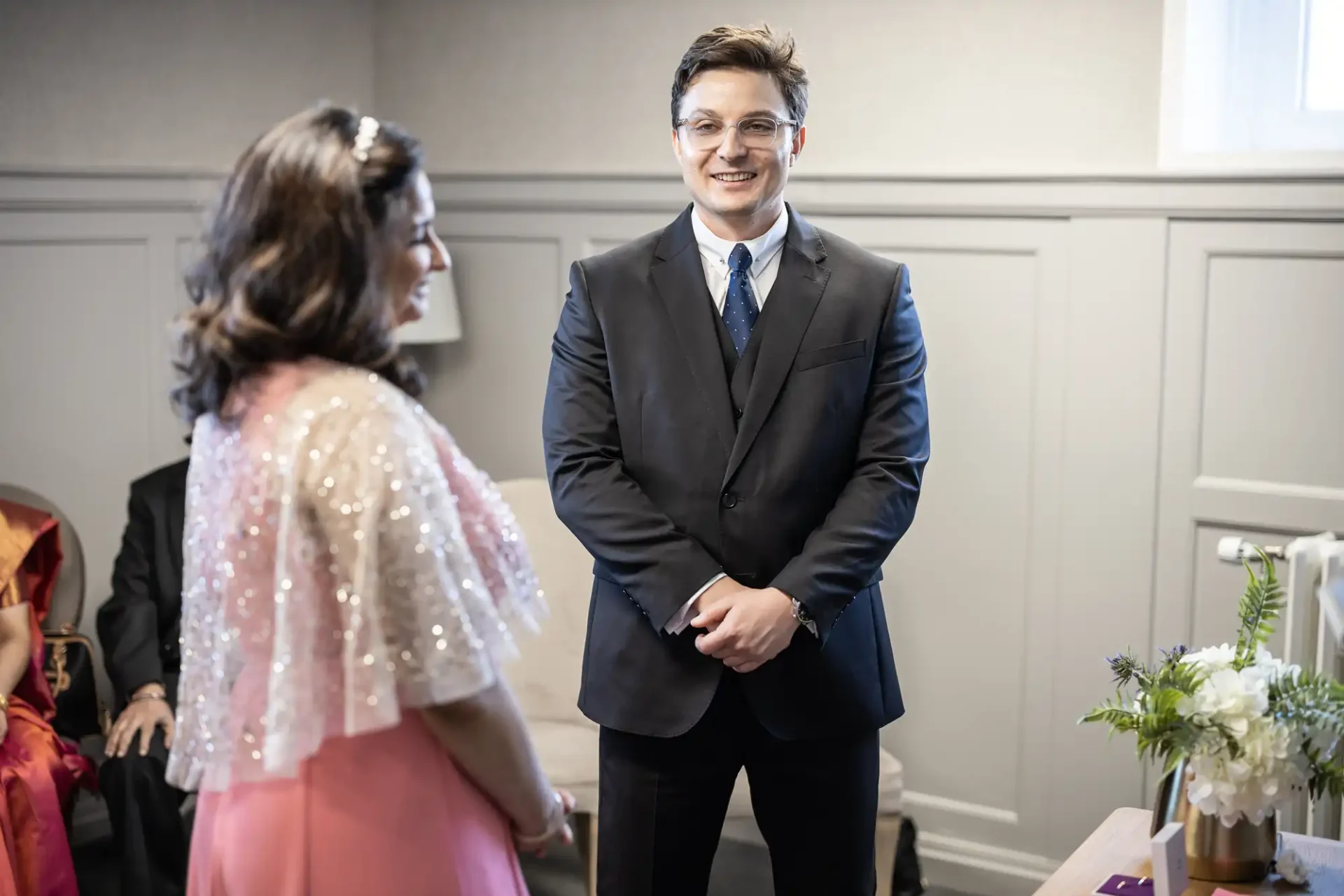 A man in a suit and a woman in a pink dress stand facing each other indoors, with two seated individuals partially visible in the background.