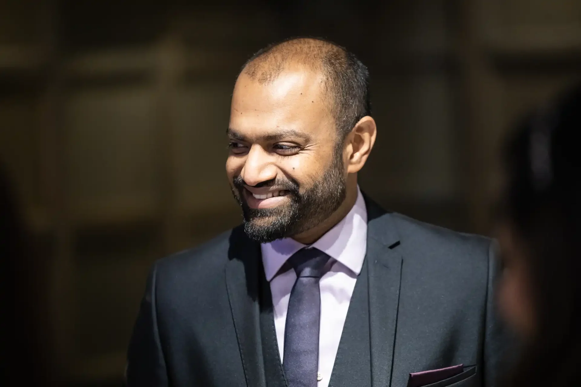 A man in a suit and tie smiles, with a blurred background.