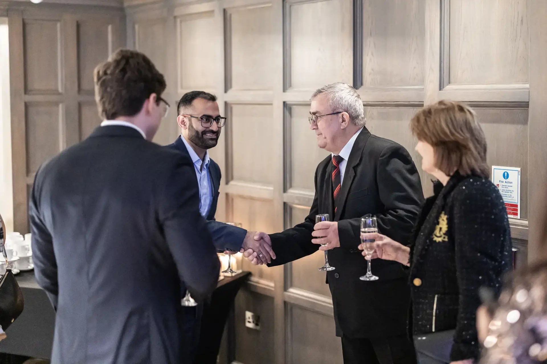 A group of people in formal attire are shaking hands and holding glasses, standing in a wood-paneled room.