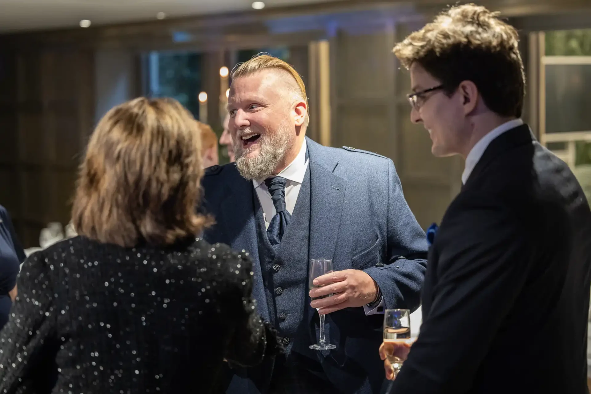 A bearded man in a blue suit smiling and holding a champagne glass, conversing with two people at an indoor gathering.