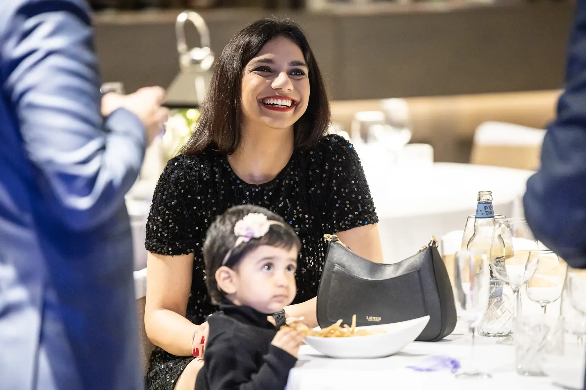 A woman in a black dress smiles at a restaurant table, with a small child in the foreground eating from a bowl.