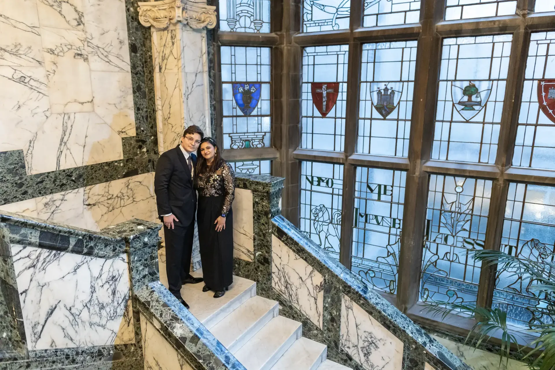 A couple stands on a marble staircase in front of large stained glass windows, adorned with colorful heraldic shields, in an ornate interior setting.