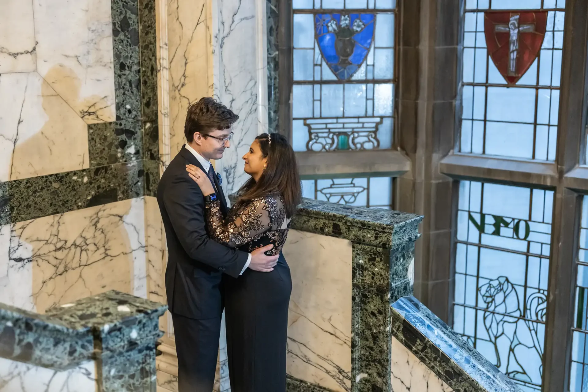 A couple embraces on a marble stairway with ornate stained glass windows in the background.