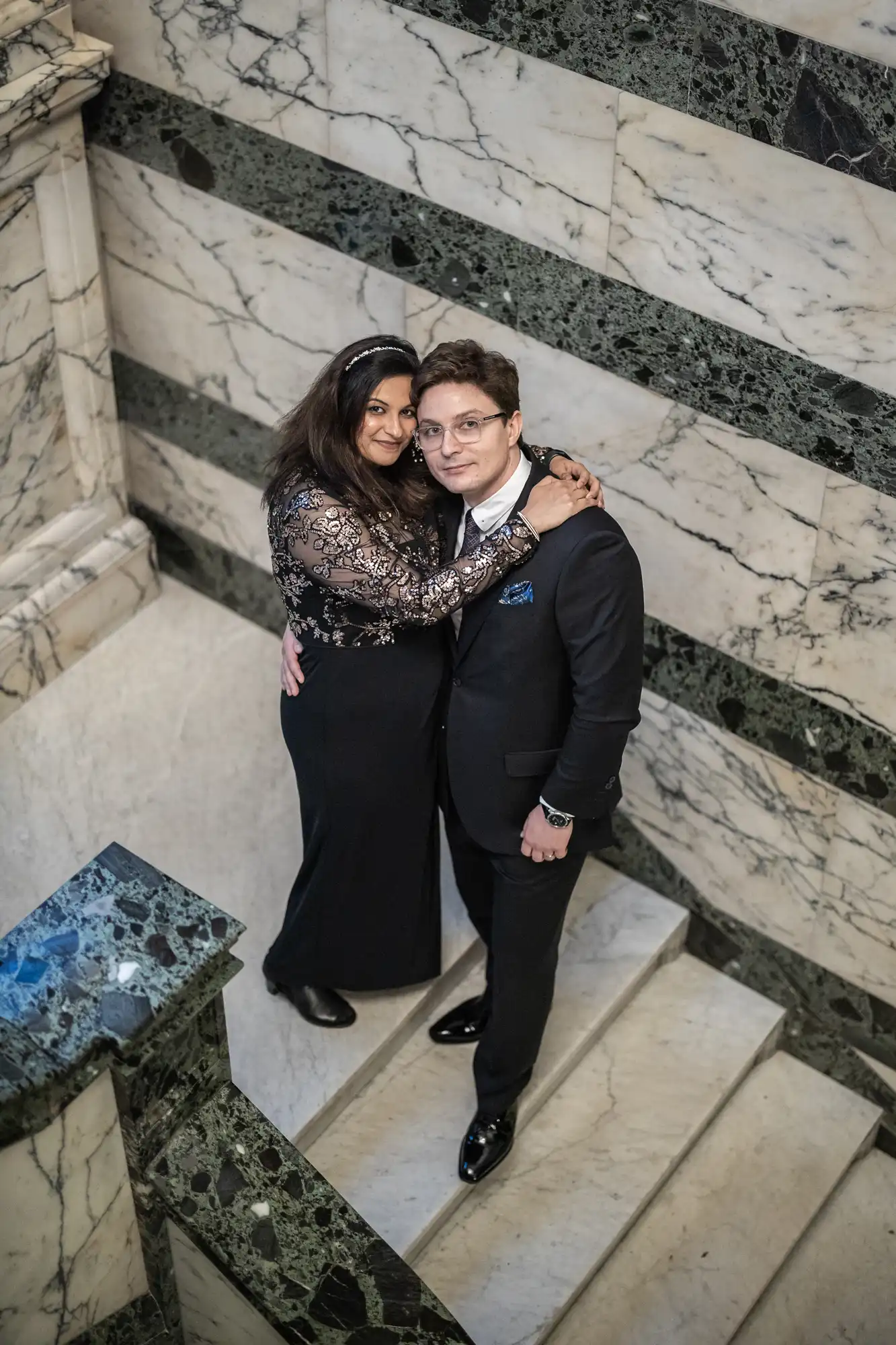 A couple poses on a marble staircase. The woman is wearing a black dress with lace and the man is in a black suit. They are embracing, looking upward. The staircase has green and white marble.