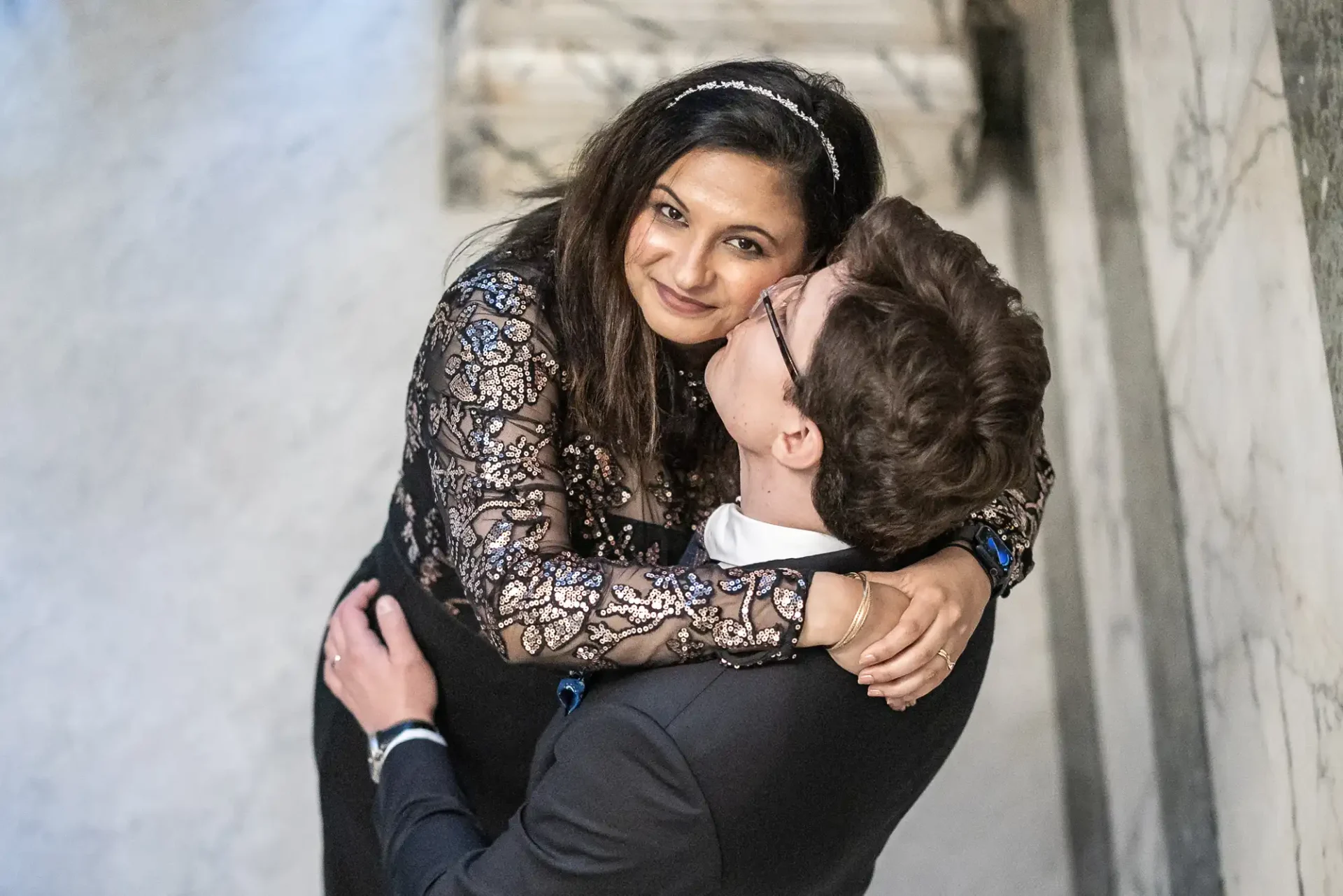 A couple embraces, with the woman smiling at the camera. She is wearing a lace dress, and the man is in a black suit, kissing her on the cheek. They are standing on a marble floor.