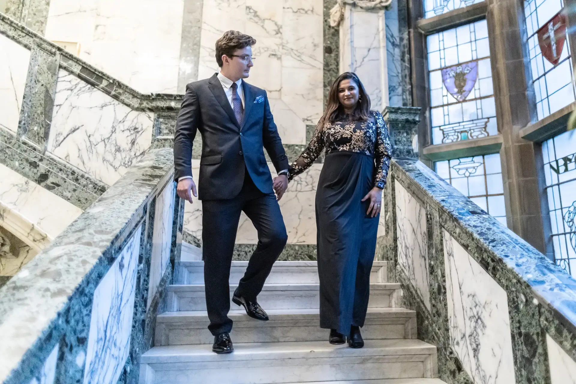 A man and woman in formal attire hold hands while descending a marble staircase in a building with stained glass windows.