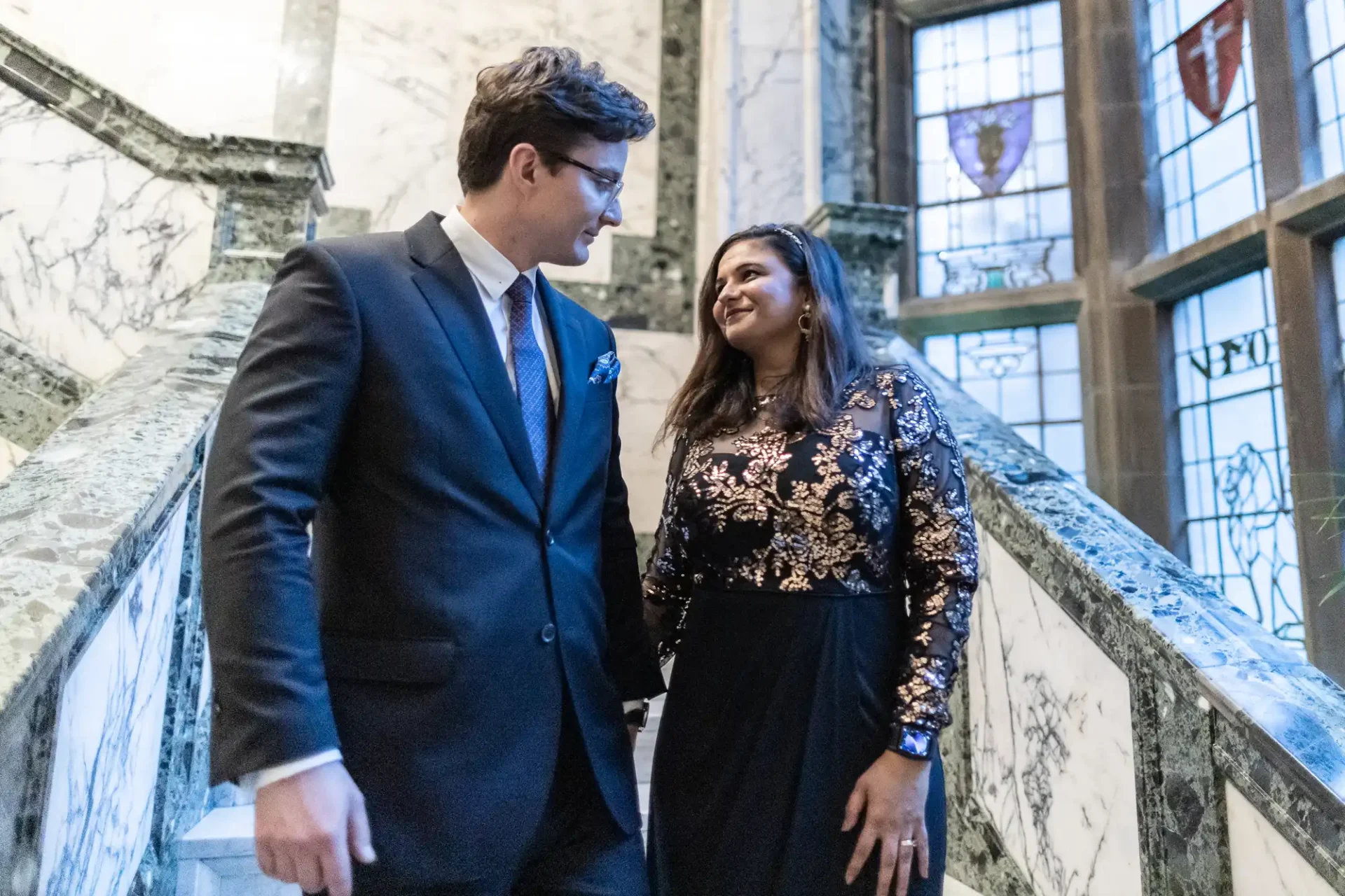 A man and woman in formal attire walk down a marble staircase, smiling at each other, with stained glass windows in the background.