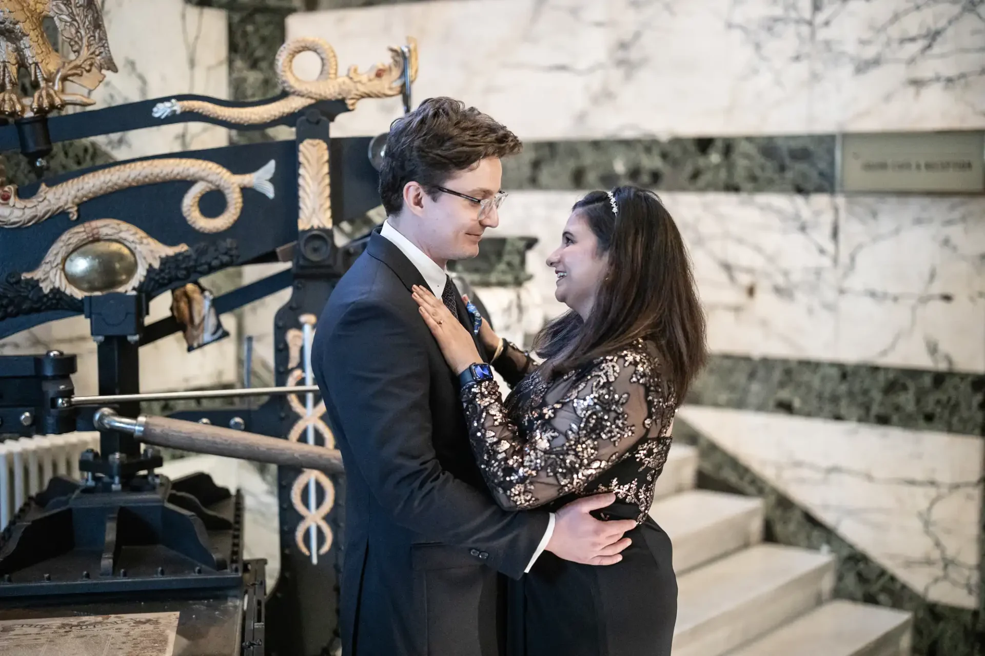 A man and woman stand embracing and smiling at each other in an ornate marble interior, with a decorative printing press in the background.