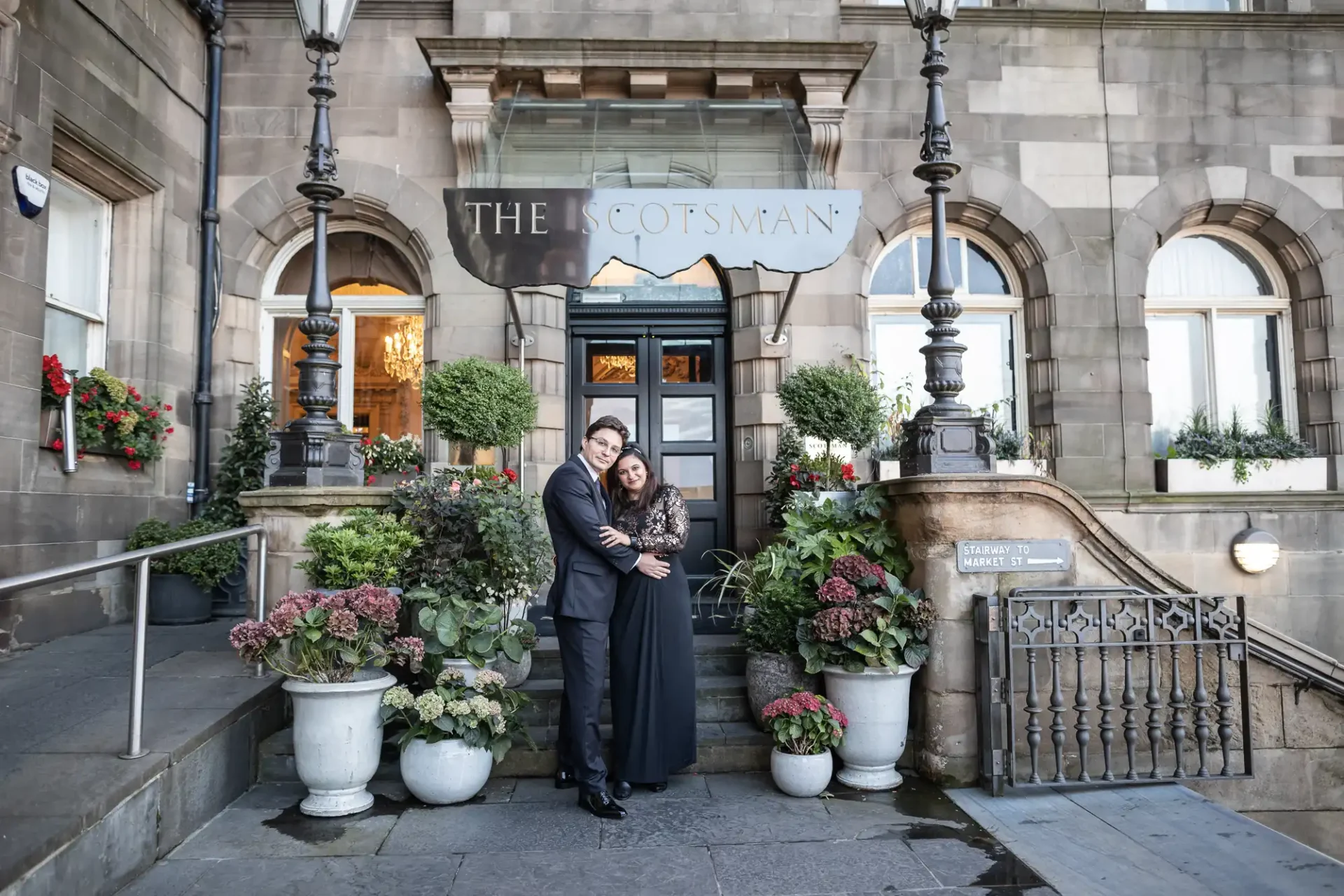 A couple embraces on the steps of The Scotsman hotel, surrounded by decorative plants and classic architecture.