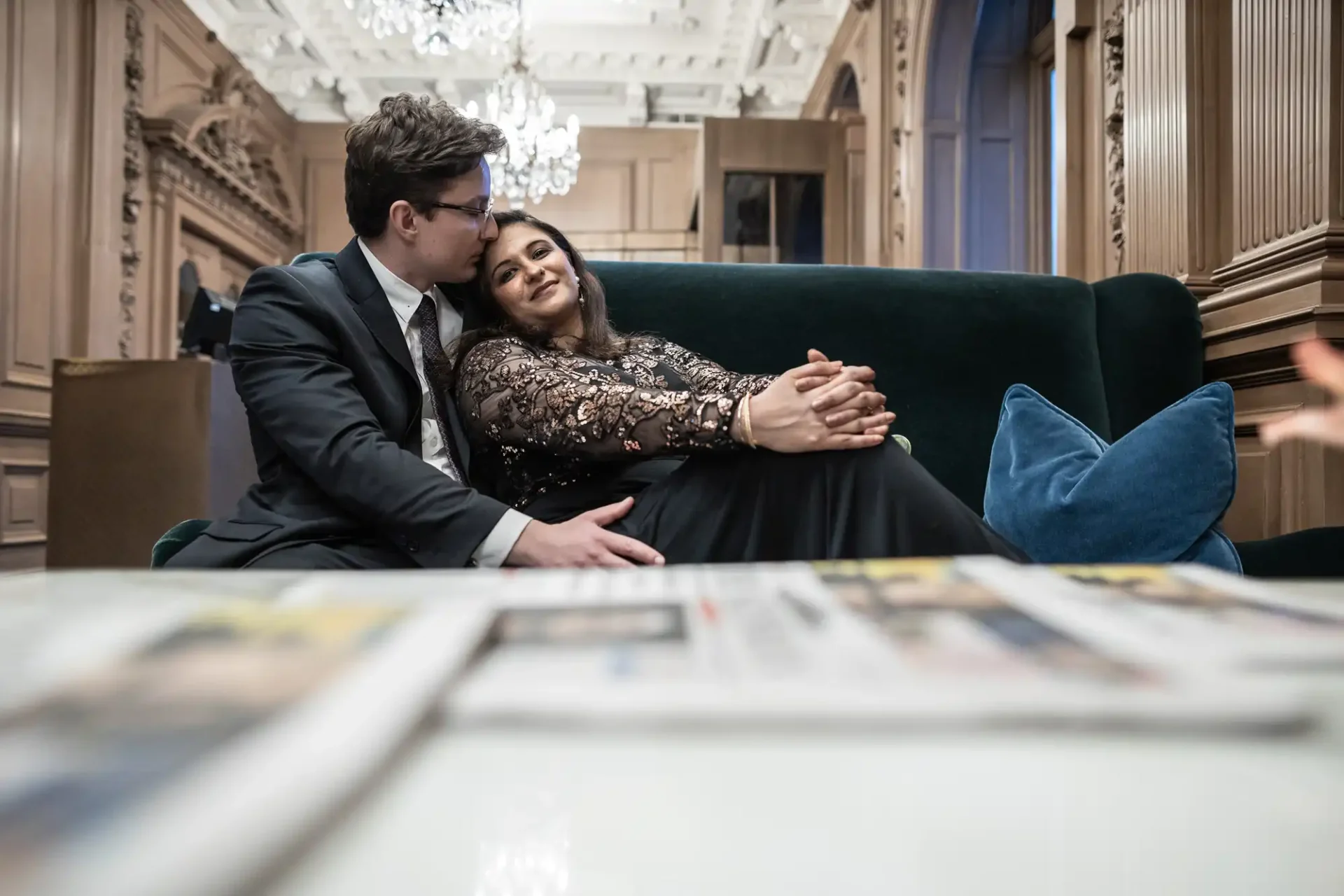 A couple sits together on a dark green couch in an elegant room. The woman rests her head on the man's shoulder. A blurred newspaper is visible in the foreground.