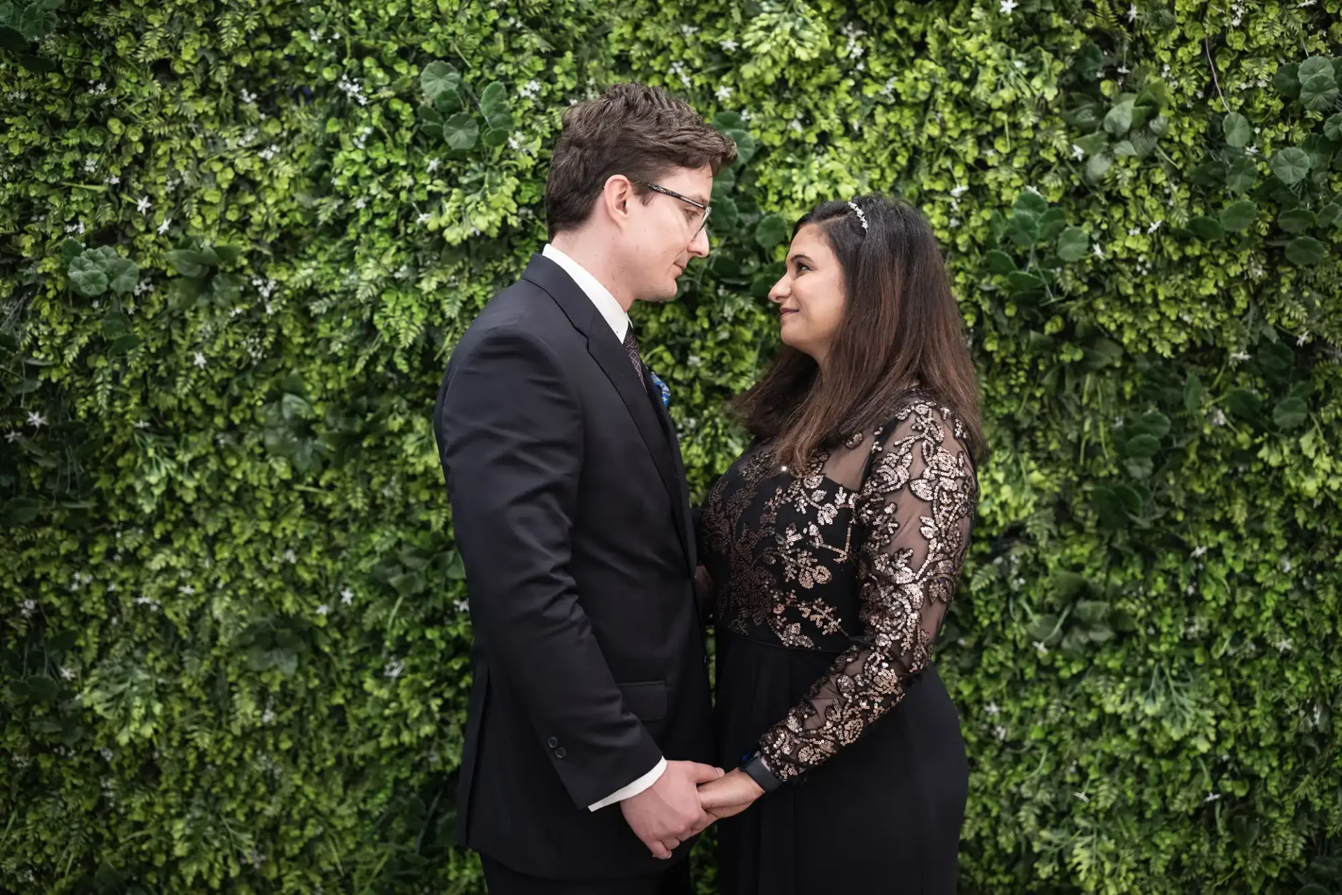 A man and woman stand facing each other, holding hands in front of a lush green foliage wall. Both wear formal attire, with the man in a suit and the woman in an embellished dress.