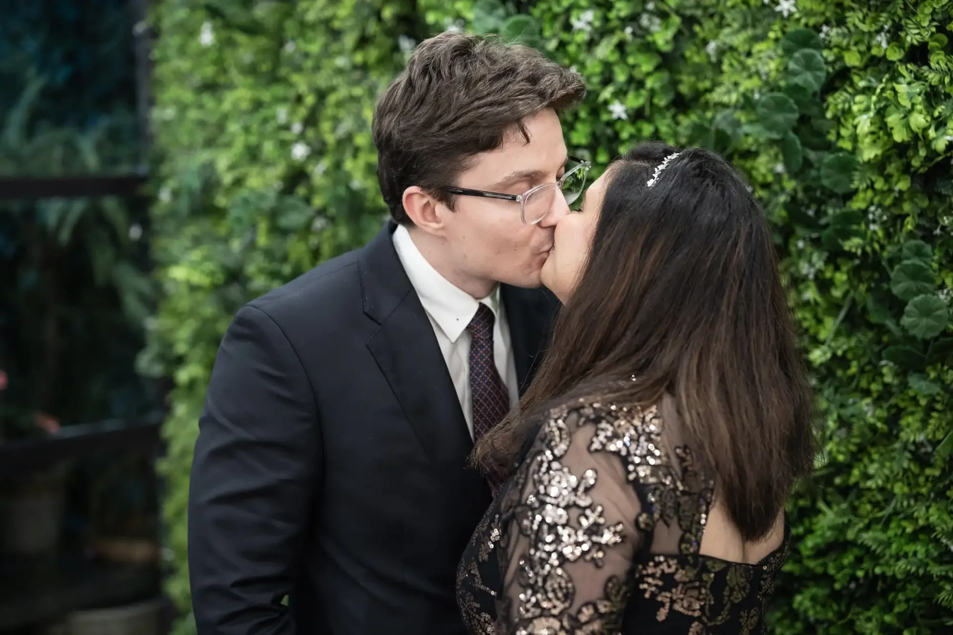 A man and woman kiss in front of a green, leafy wall. The man wears glasses and a suit, while the woman wears an ornate, lace-patterned dress.