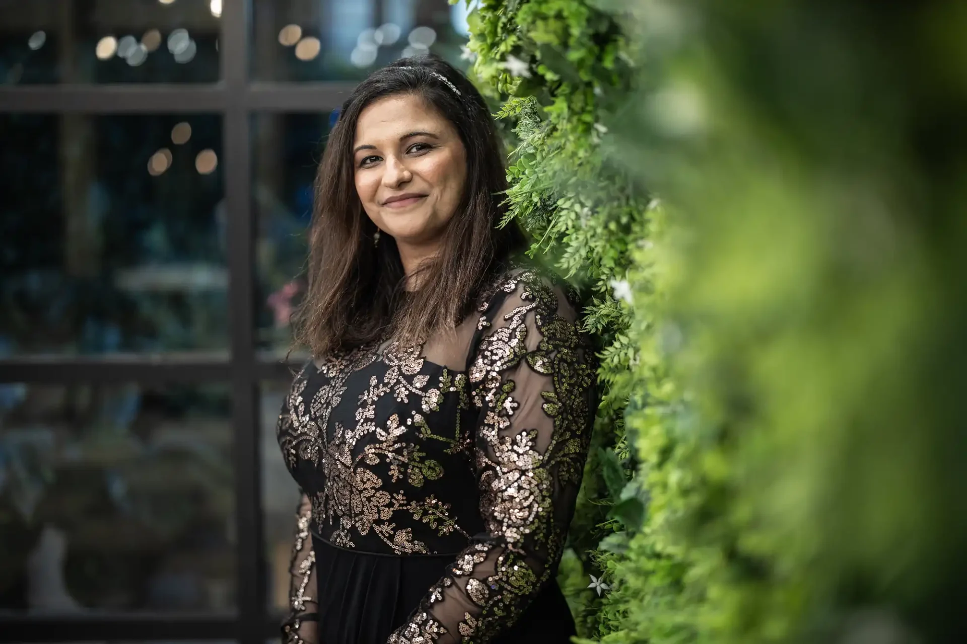 A woman in a black and gold dress stands smiling against a green leafy wall with glass windows in the background.