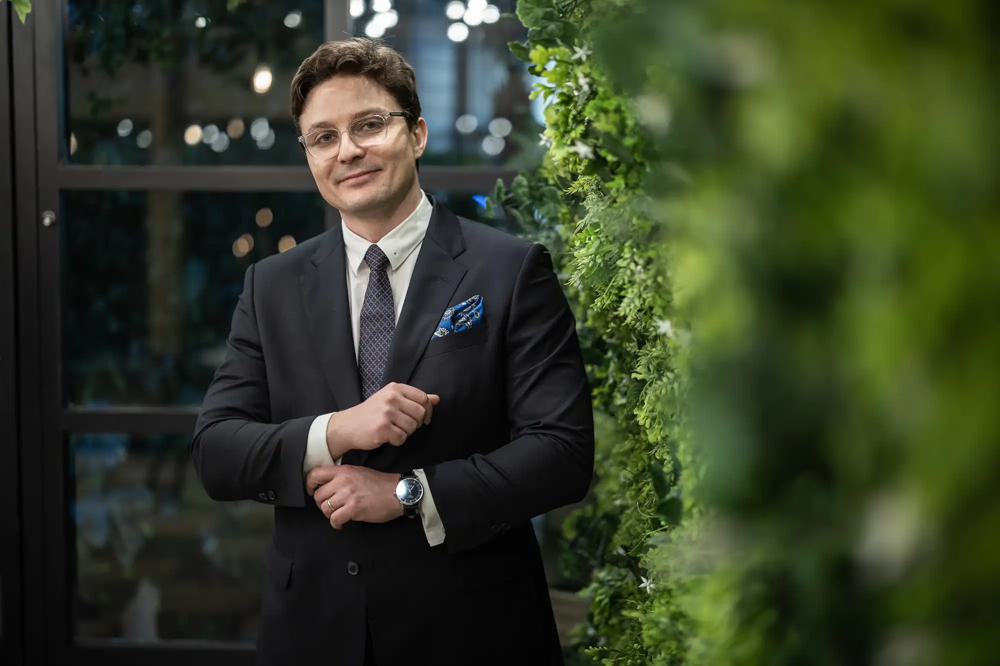 A man in a suit, wearing glasses and a watch, adjusts his cuff while standing next to a wall of green foliage indoors.