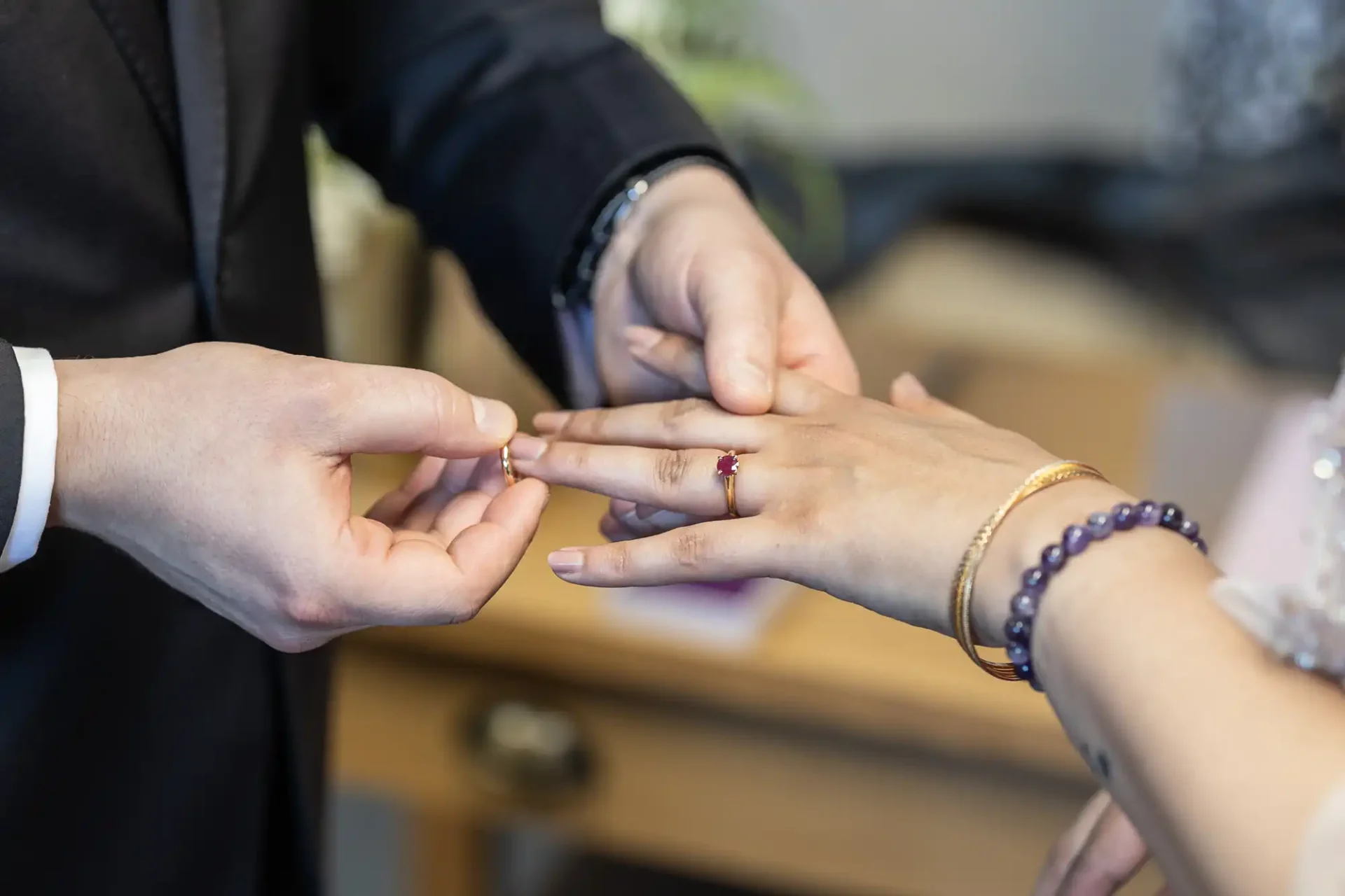 A person places a ring on another's finger, with both wearing formal attire and various bracelets visible.