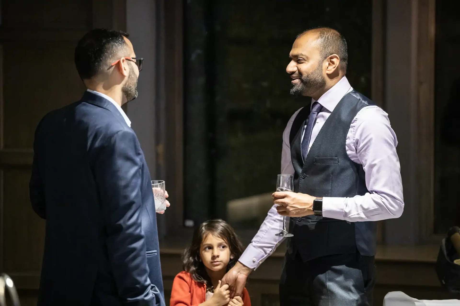 Two men in conversation holding drinks, with a young girl seated in front of them at a formal event.