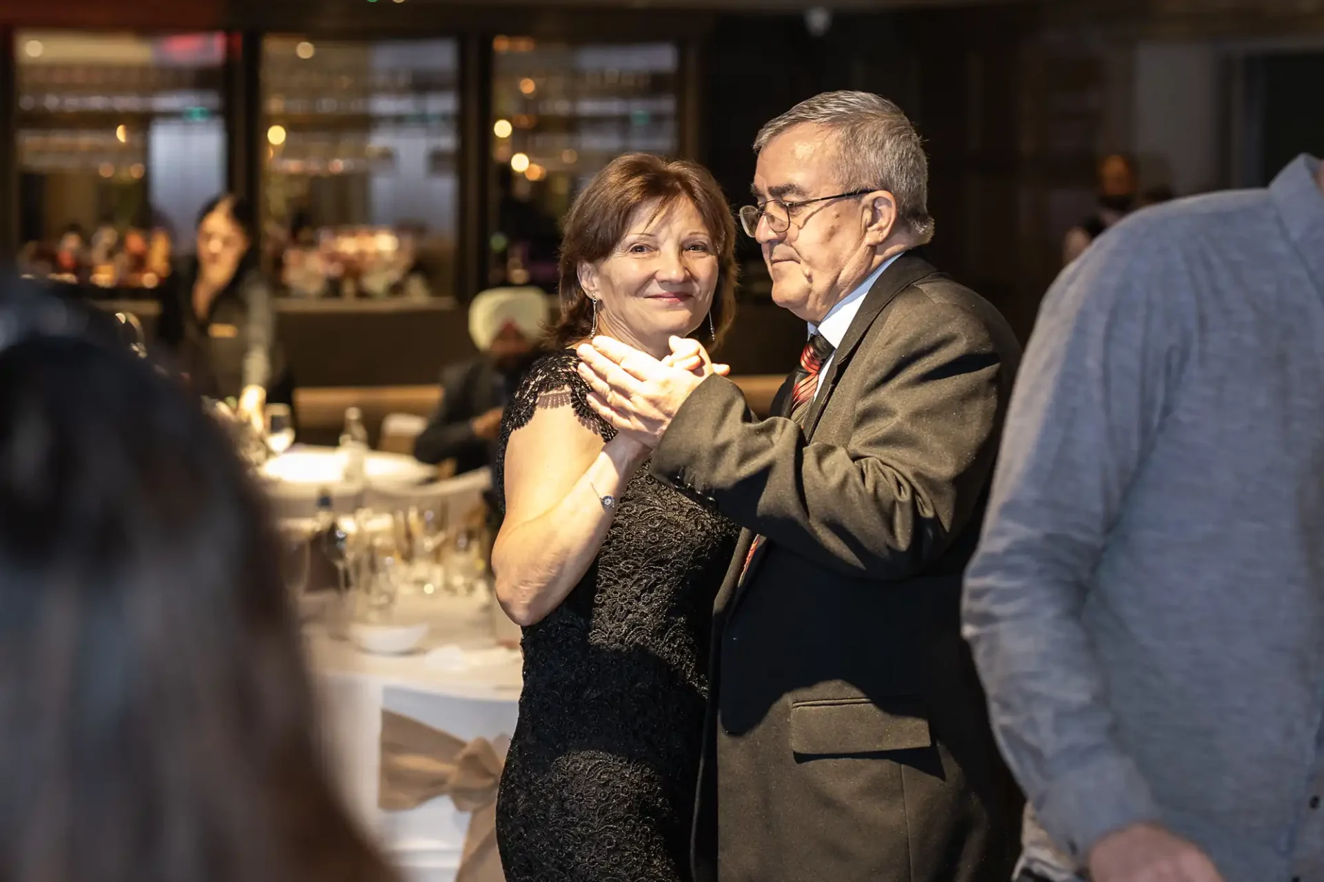 An older couple dances closely at a formal indoor event, with tables and guests visible in the background.