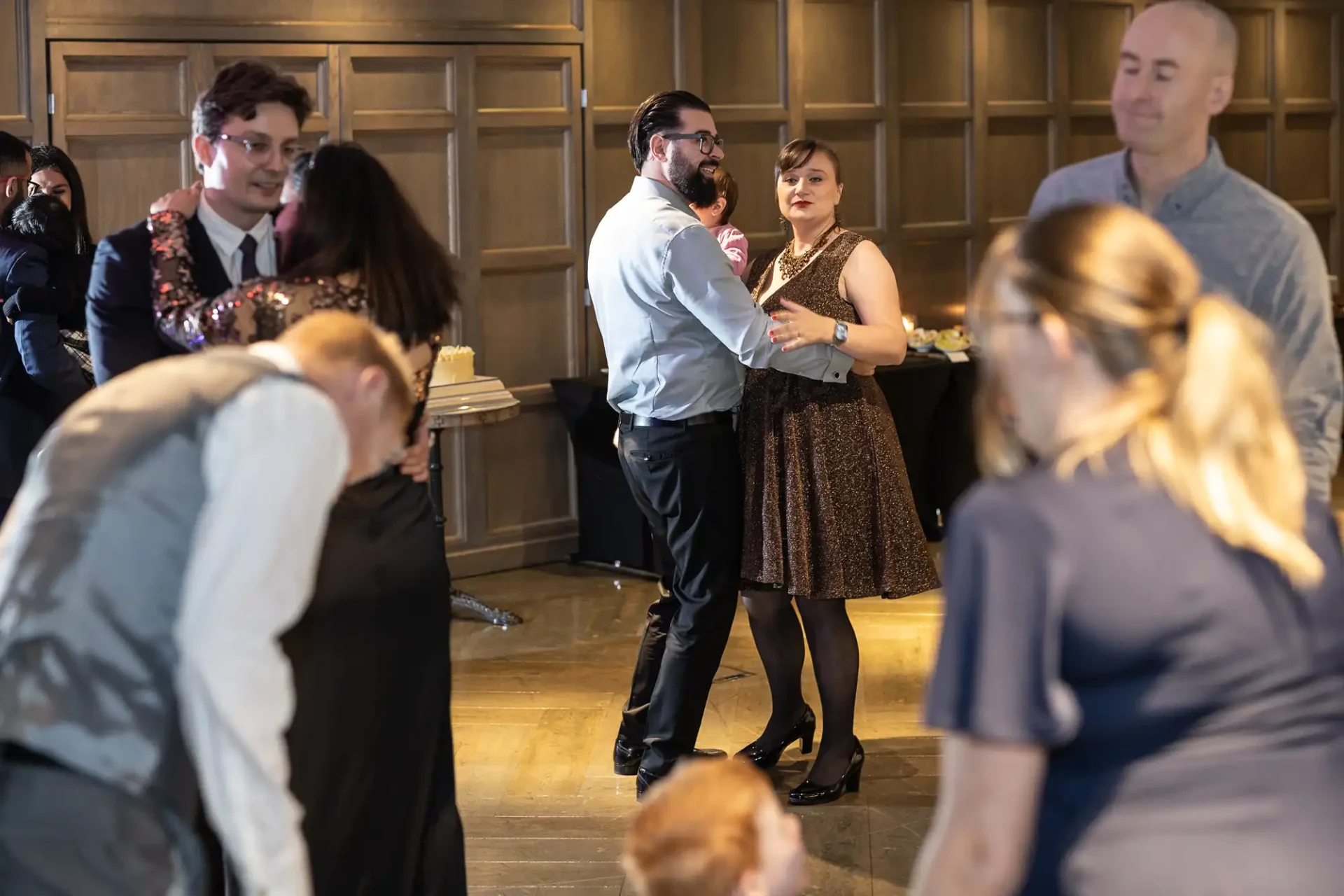 People dancing in a room with wooden panel walls. A few couples are moving together, while others stand nearby. Tables with food are visible in the background.