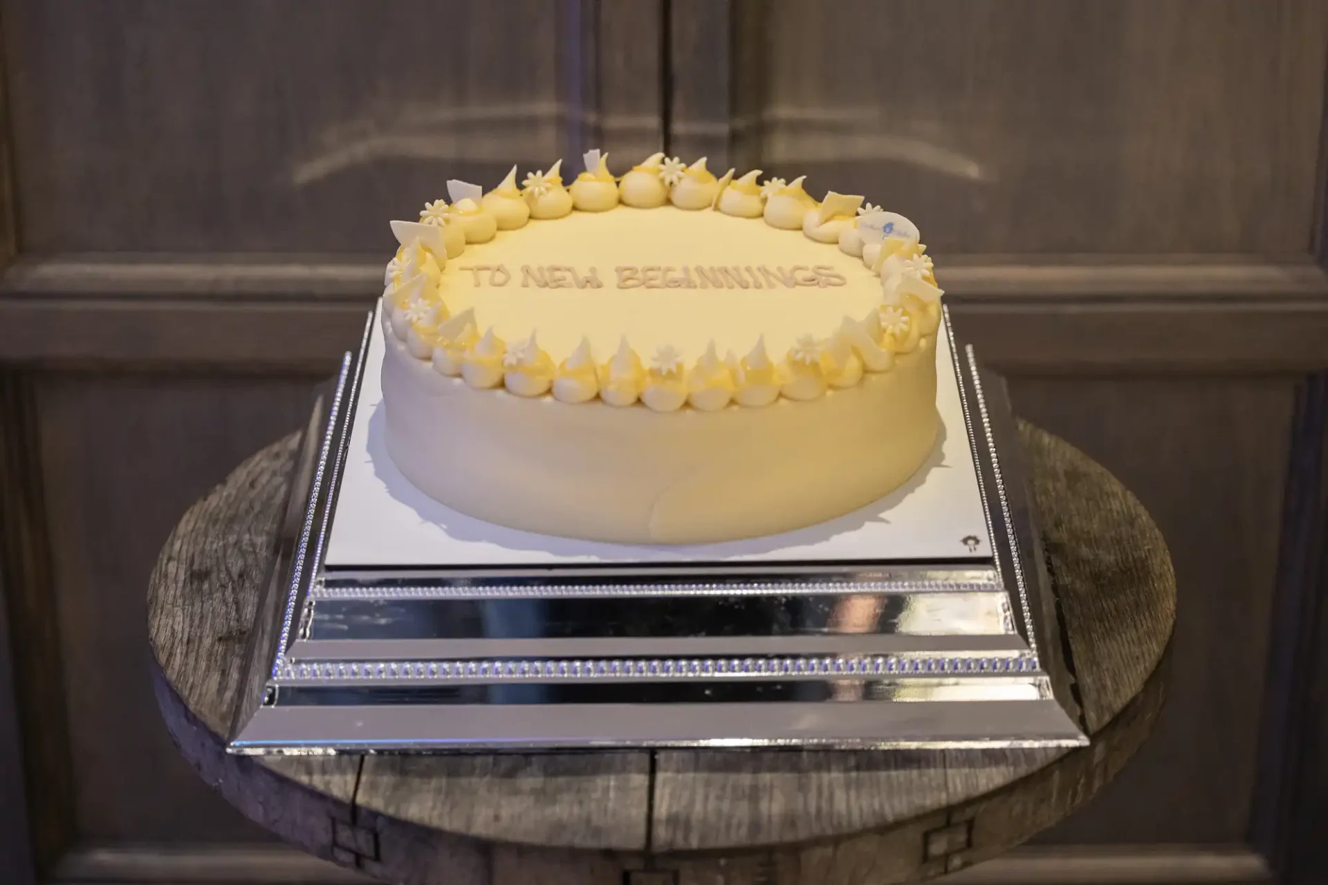 A white cake with "To New Beginnings" written on top, decorated with frosting peaks. The cake is placed on a mirrored stand on a wooden surface.