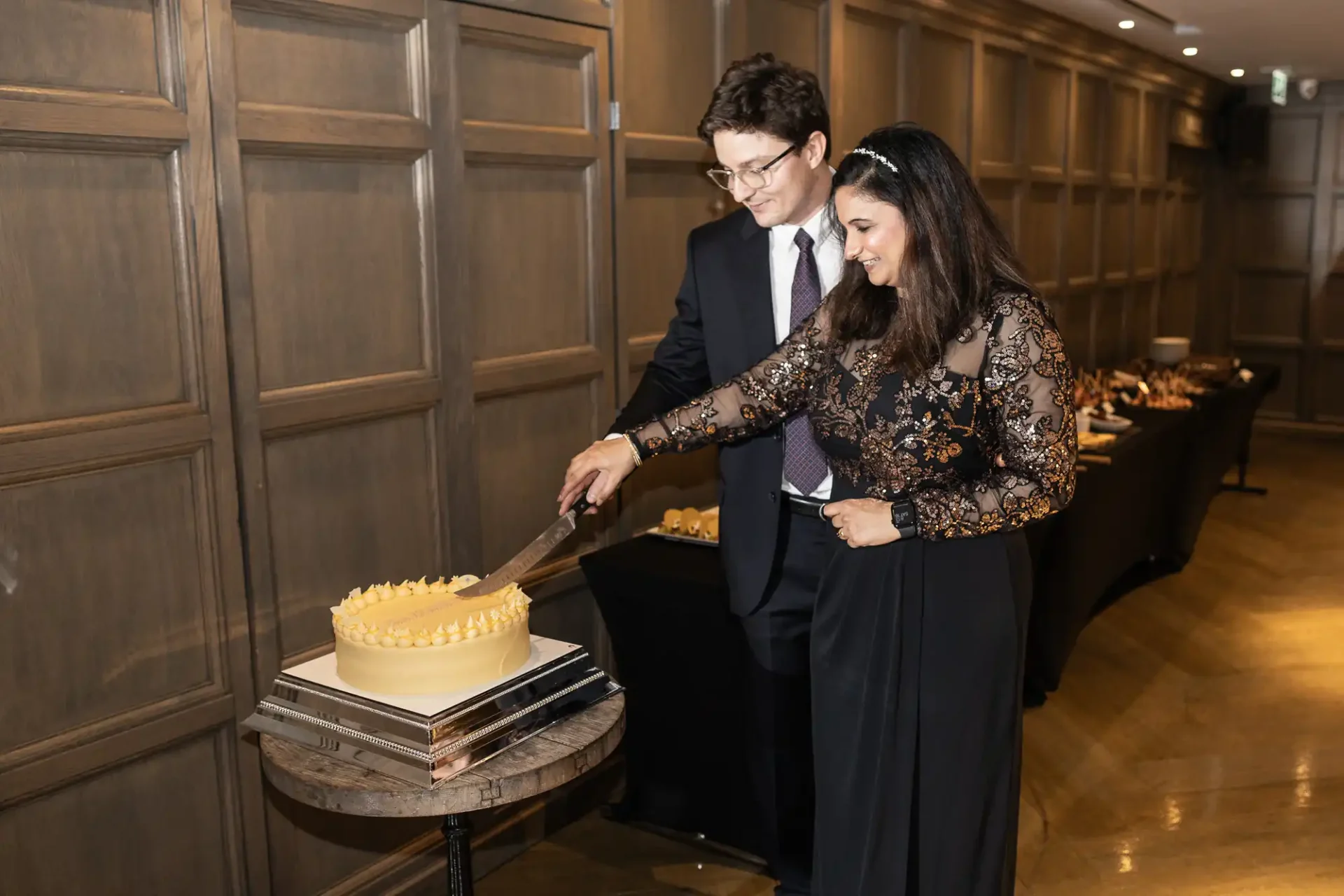 A man and woman, both dressed formally, are cutting a round cake with yellow frosting on a small table in a wood-paneled room.