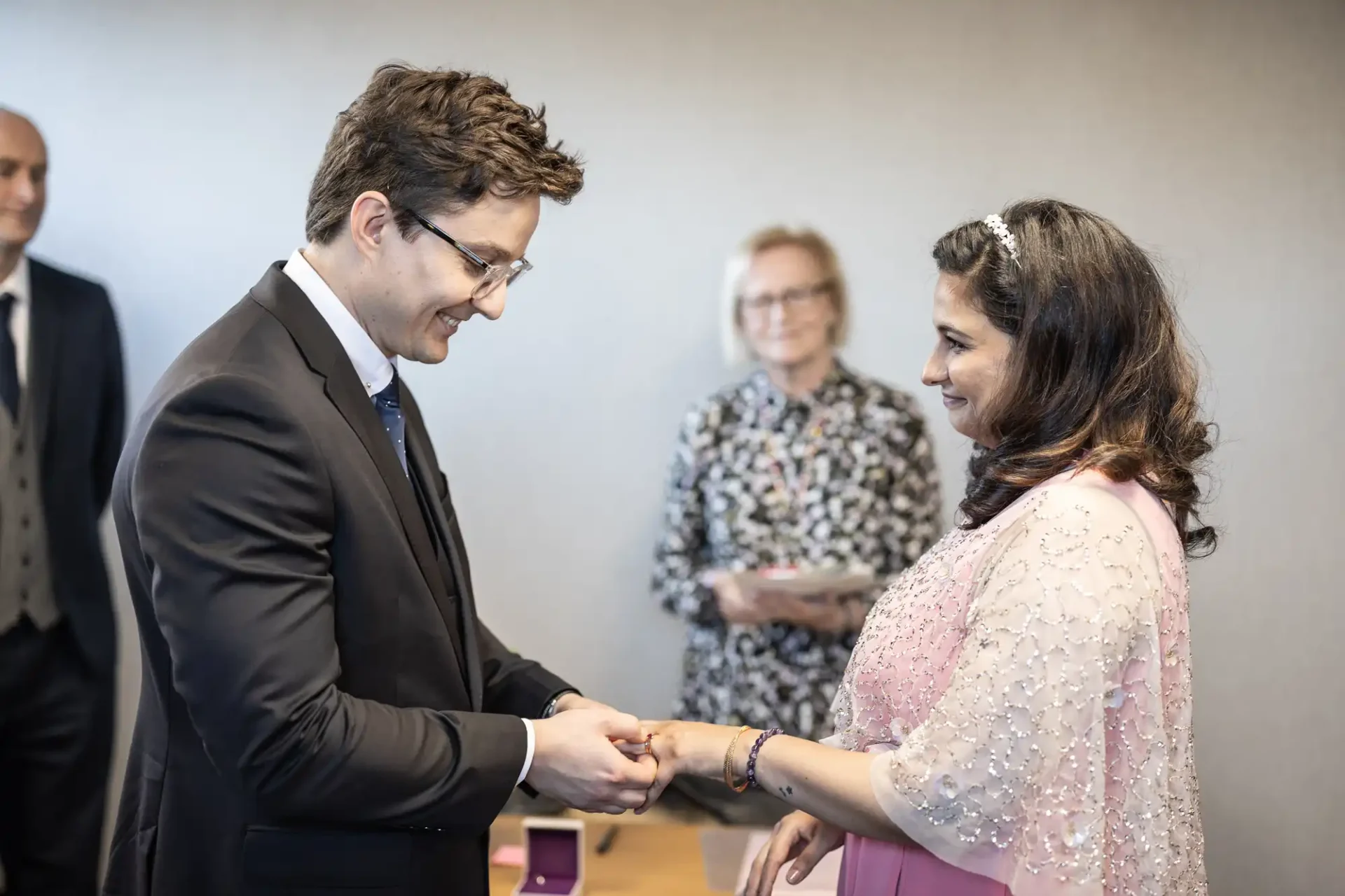 A man in a suit places a ring on a woman's finger during a ceremony. A woman in the background observes while holding a clipboard.