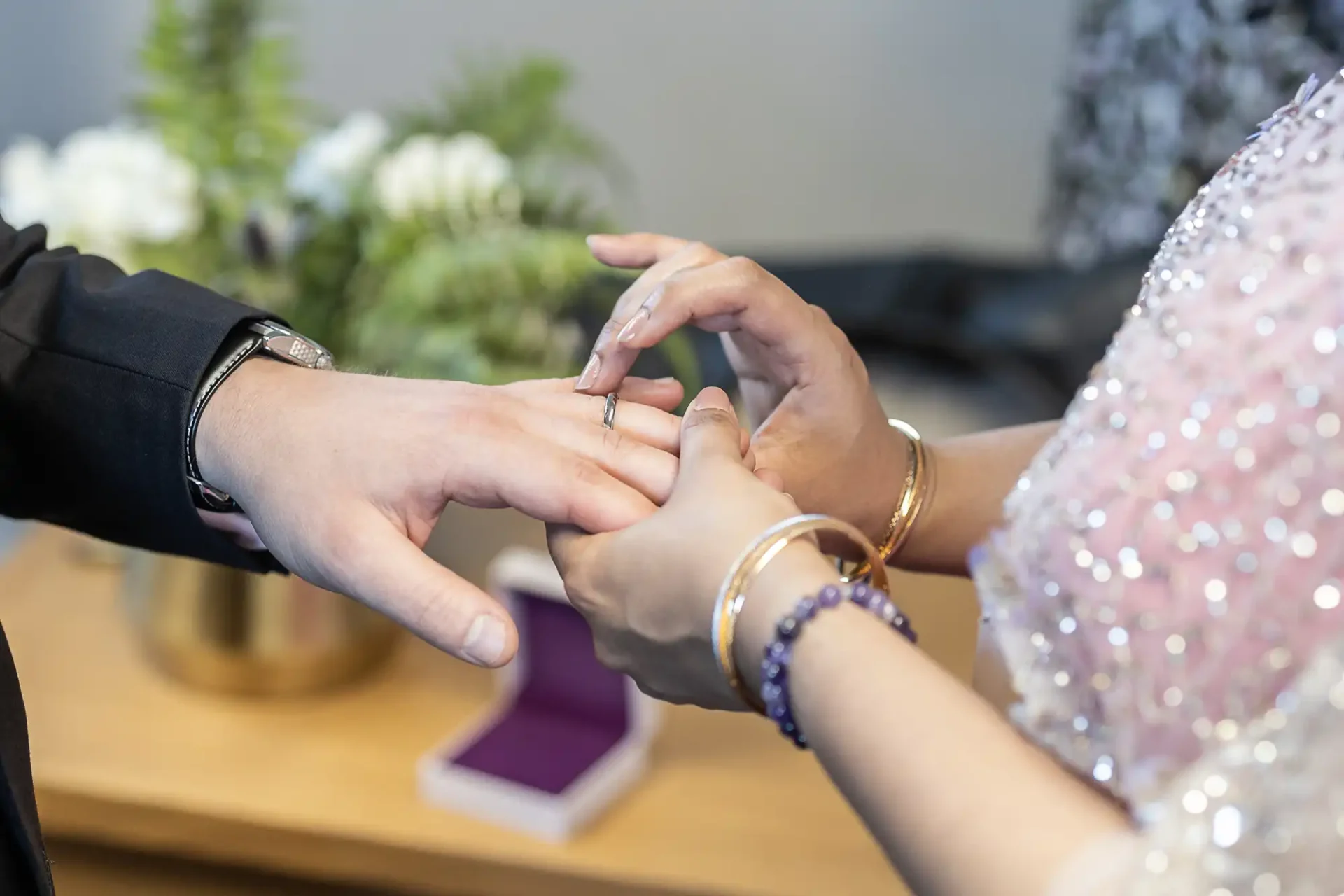 A person places a ring on another's finger in a ceremony setting, with a flower arrangement and an open box in the background.