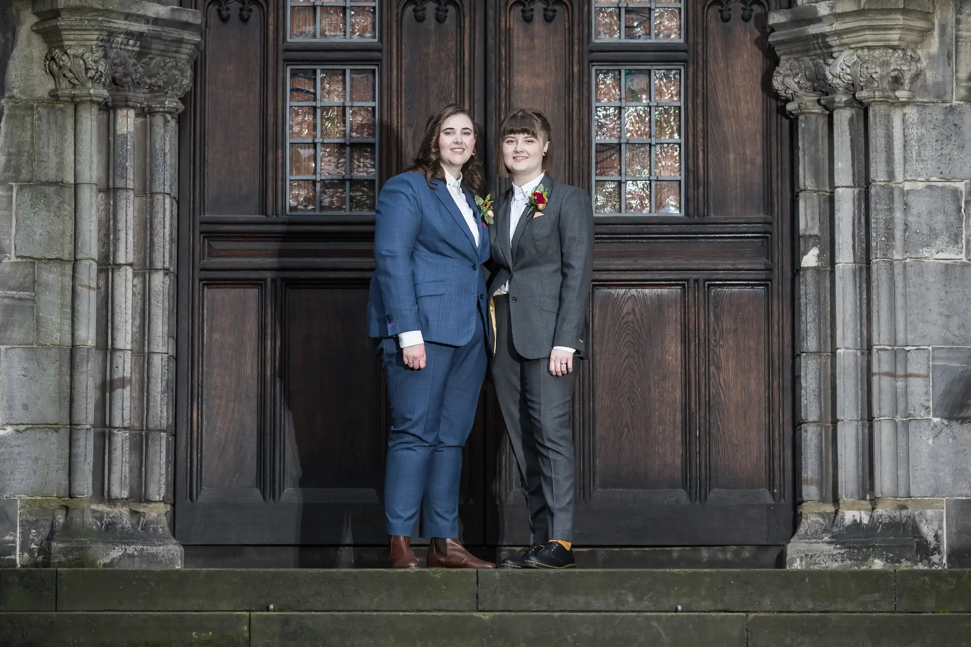 Two newlywed ladies in suits stand side by side on steps in front of a large wooden door with ornate stone columns on either side. Both are smiling and looking at the camera.