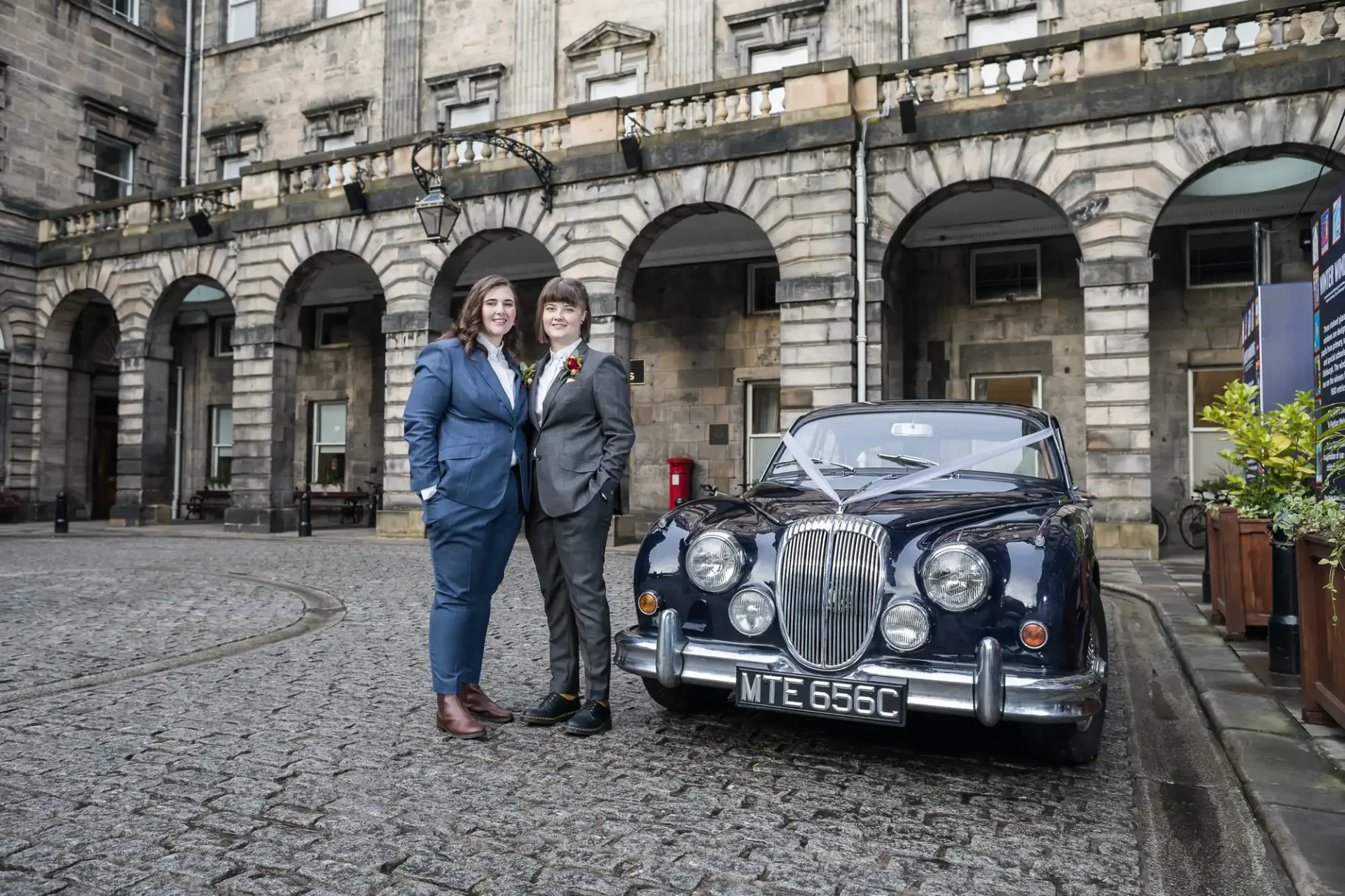 Two newlywed ladies in wedding suit attire stand side by side on a cobblestone street, in front of an ornate building and a vintage car with the license plate MTE 656C.