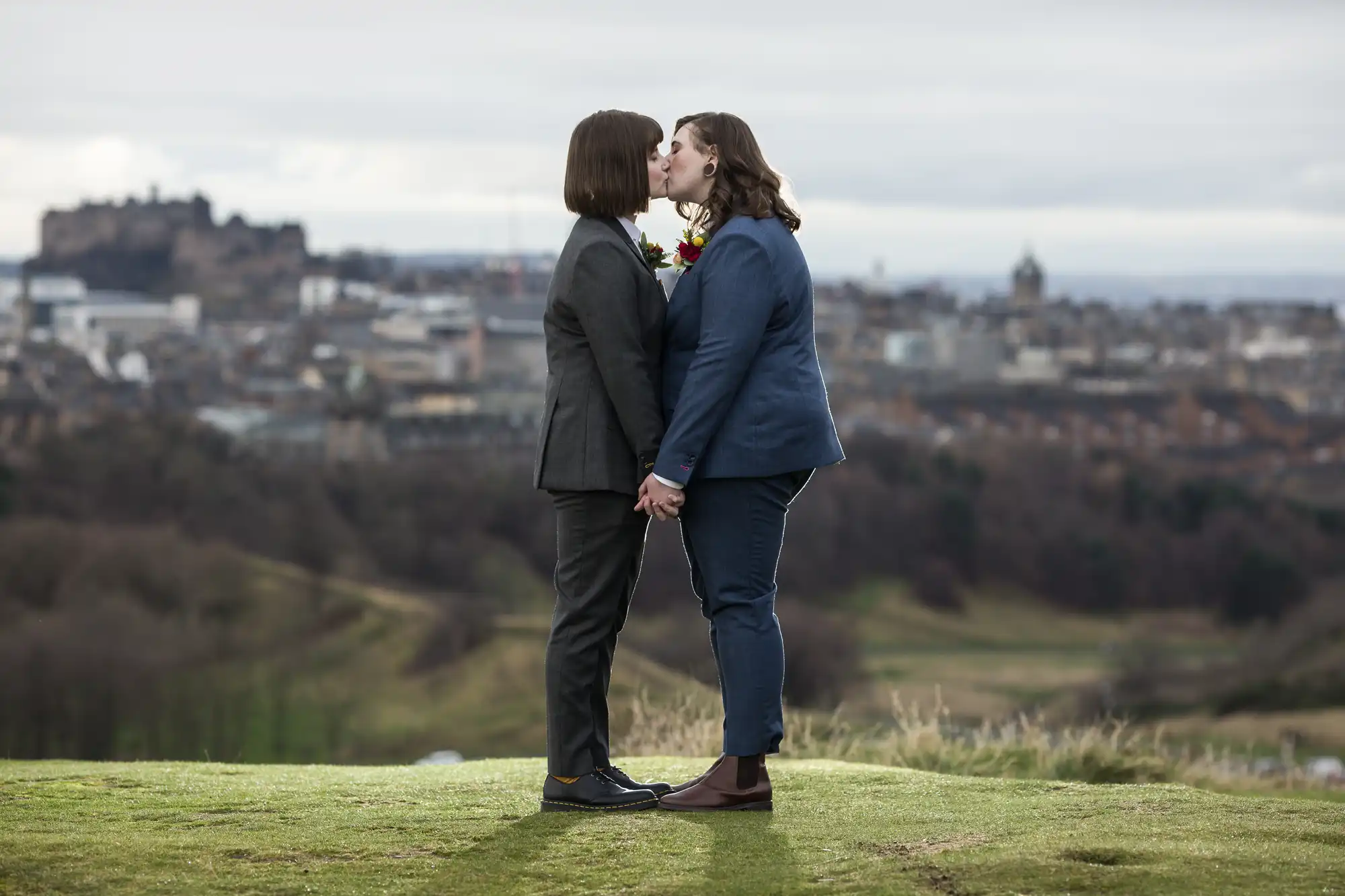Two individuals in suits share a kiss while holding hands on a grassy hill, with a cityscape and a castle visible in the background.
