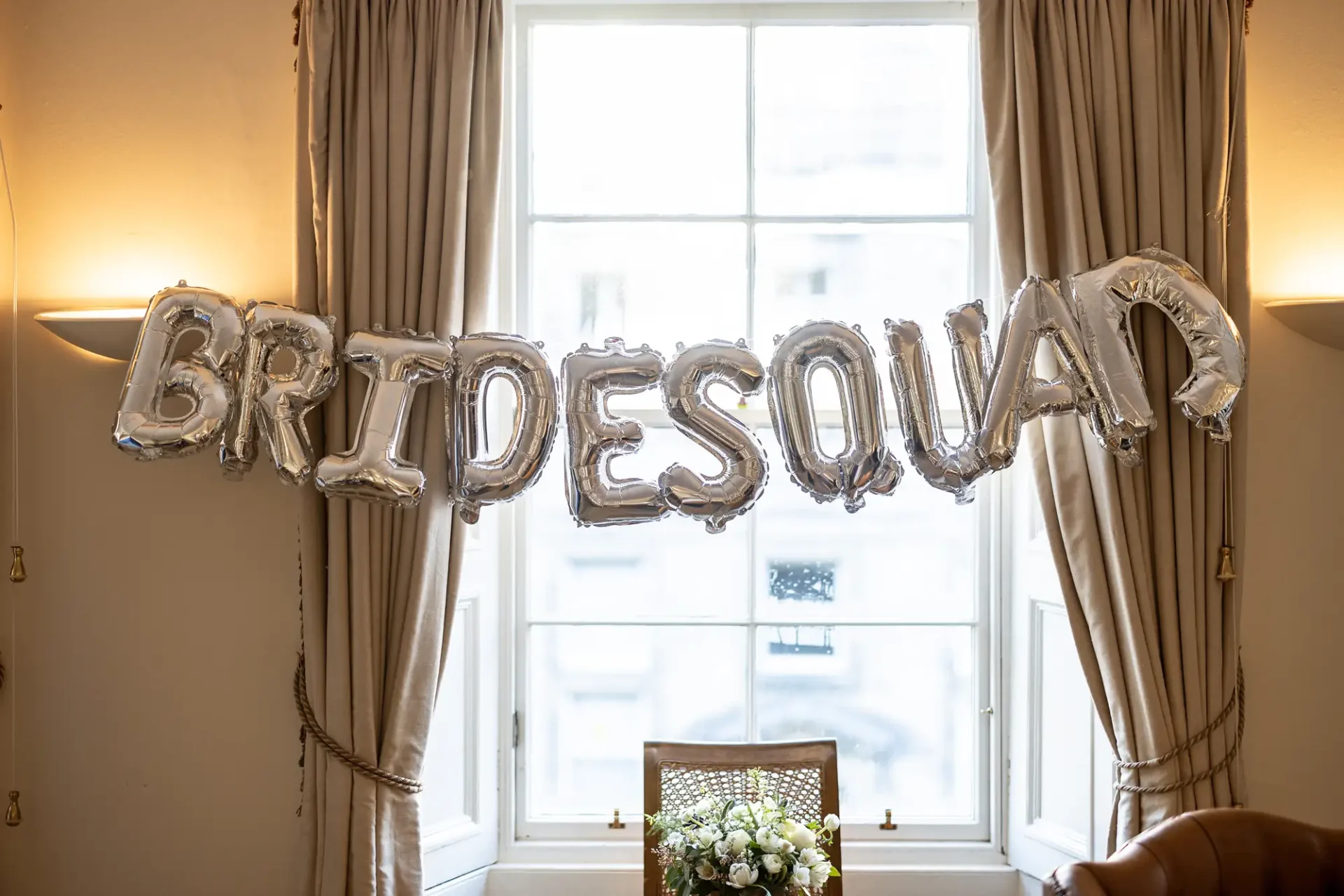 Silver "bride squad" balloon letters hanging in front of a window with curtains in a room with warm lighting.