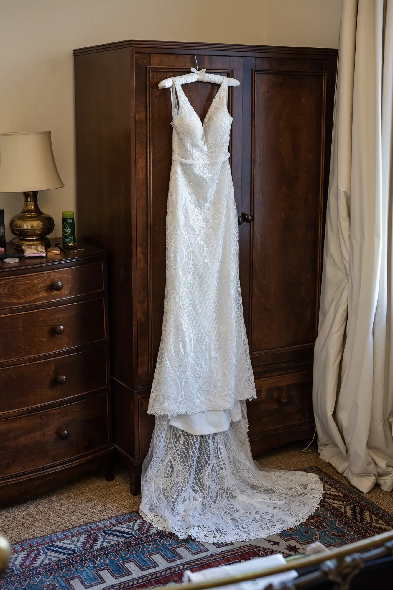 A white wedding dress with lace detailing hangs in front of a dark wood wardrobe in a bedroom setting.