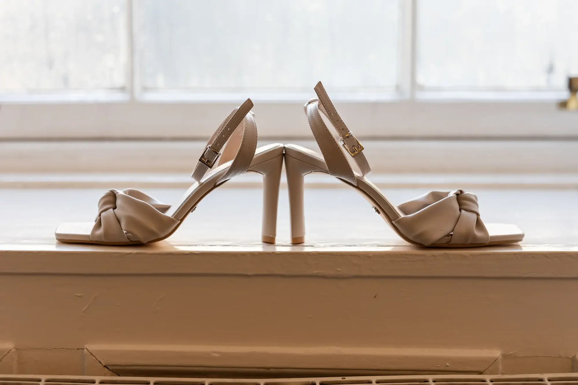A pair of elegant beige high-heeled shoes displayed on a windowsill against a softly lit background.