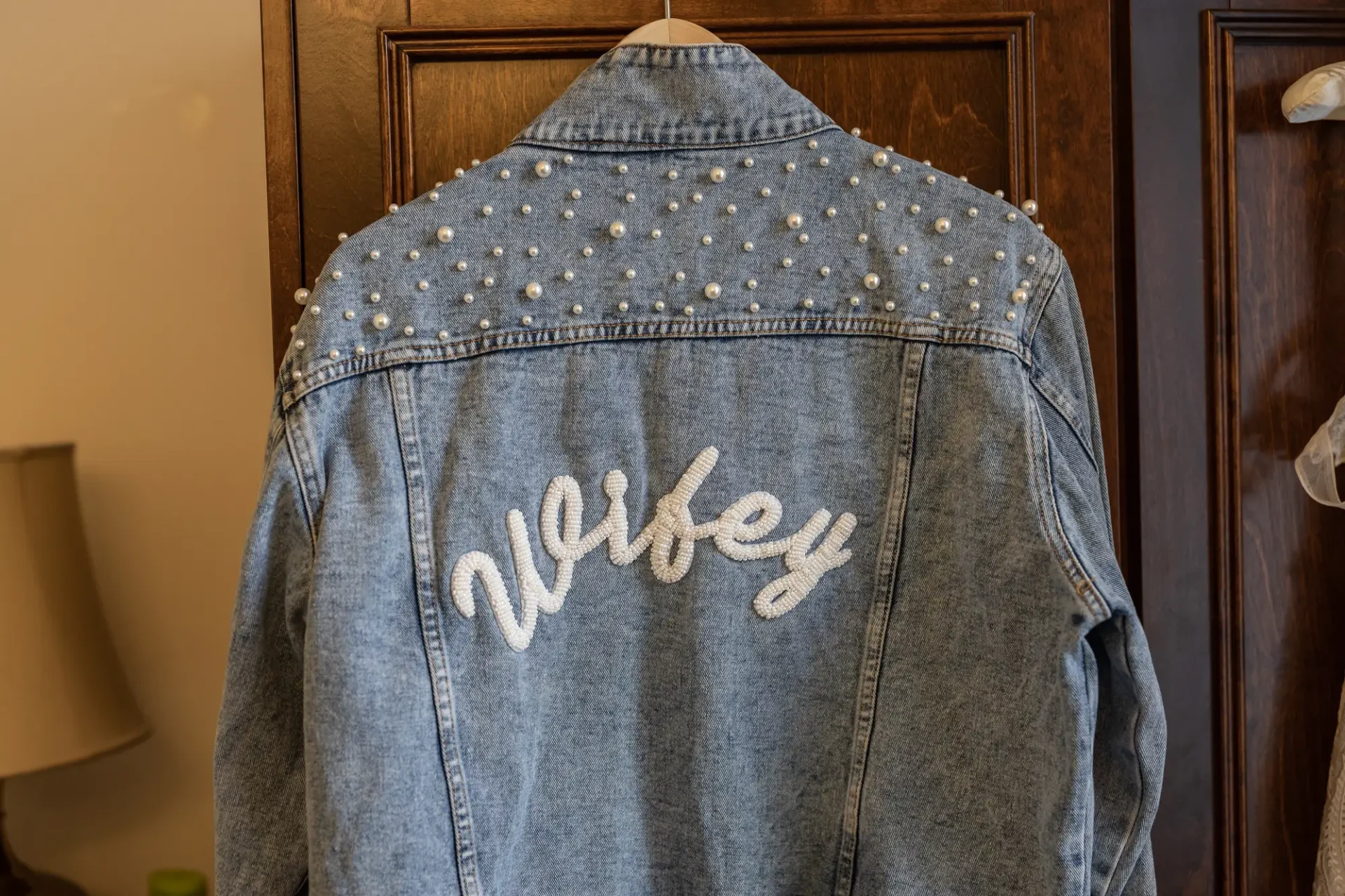 A denim jacket with the word "wifey" embroidered in white script and adorned with pearl embellishments, hanging on a wooden door.