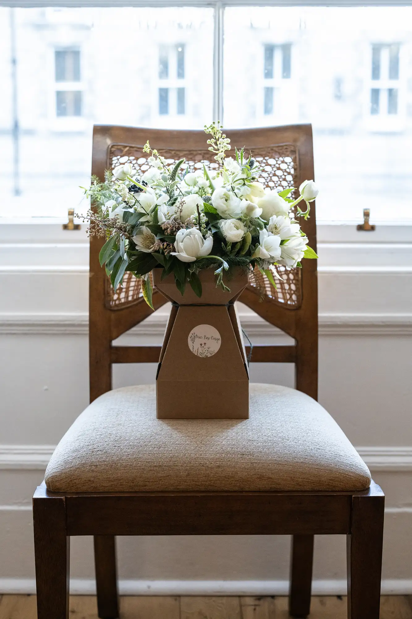 A floral arrangement in a paper bag sits on a chair by a window, featuring predominantly white flowers and greenery.