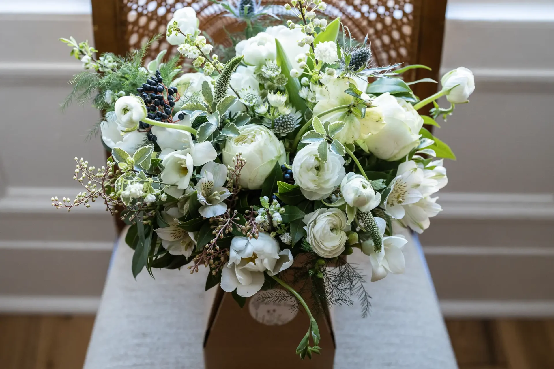 A floral bouquet with white roses, green thistles, and assorted greenery rests on a person's lap against a chair background.