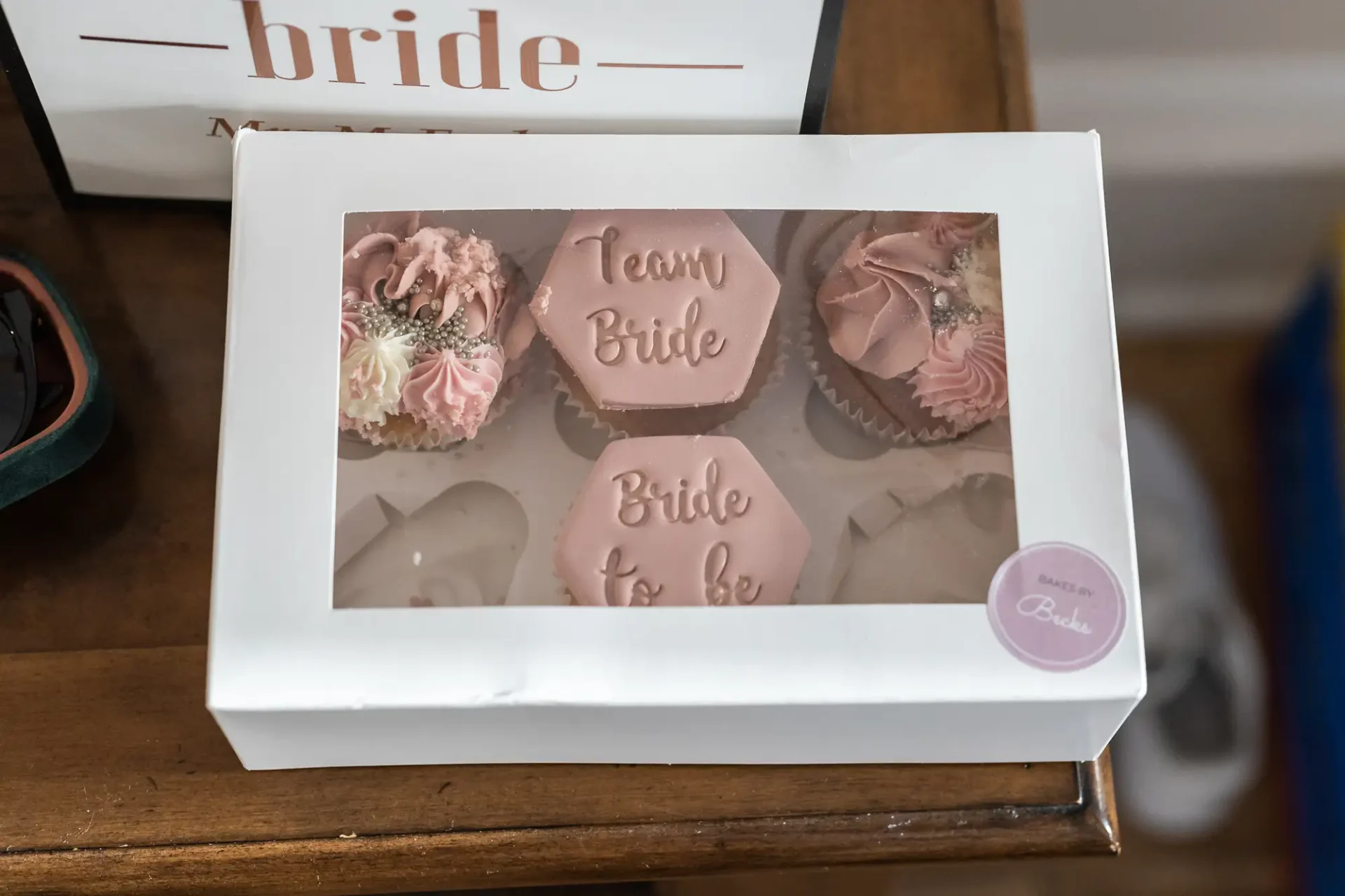 A box of pink and white themed cupcakes with "team bride" and "bride to be" decorations, placed on a wooden surface next to a "bride" sign.