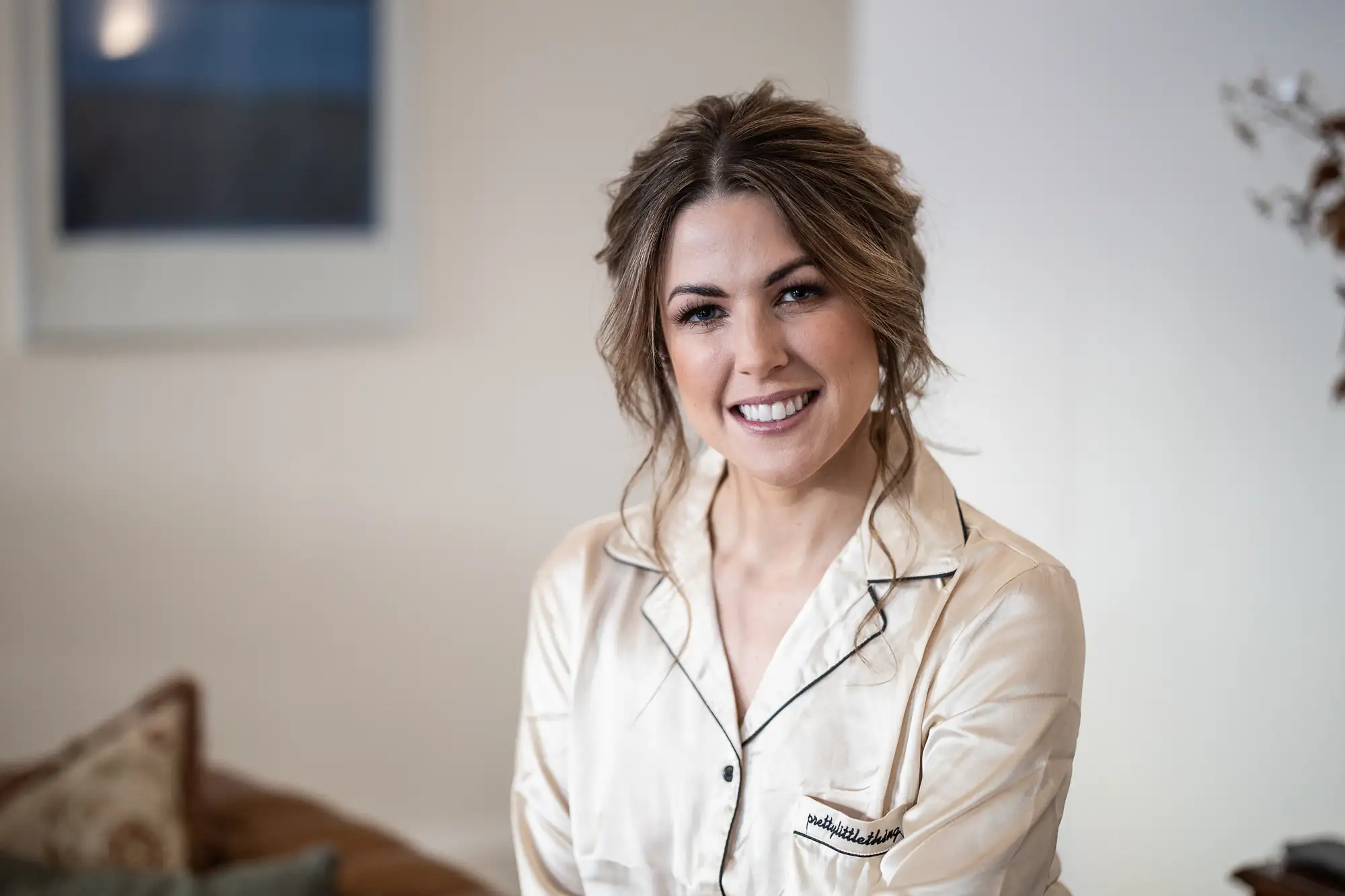 A smiling woman wearing a beige shirt with a name tag sits in a warmly lit room with a soft-focus background.