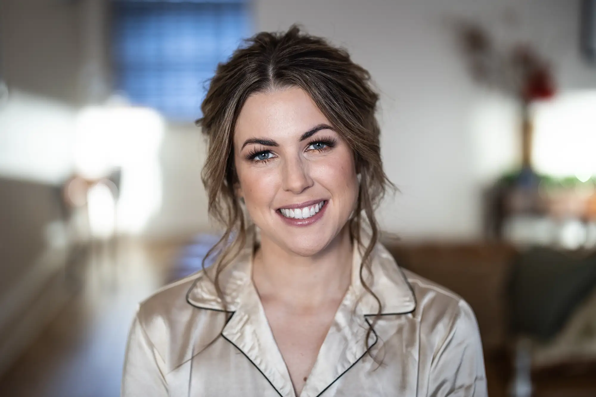 Portrait of a smiling woman with brown hair, wearing a light beige shirt, indoors with a softly blurred background.