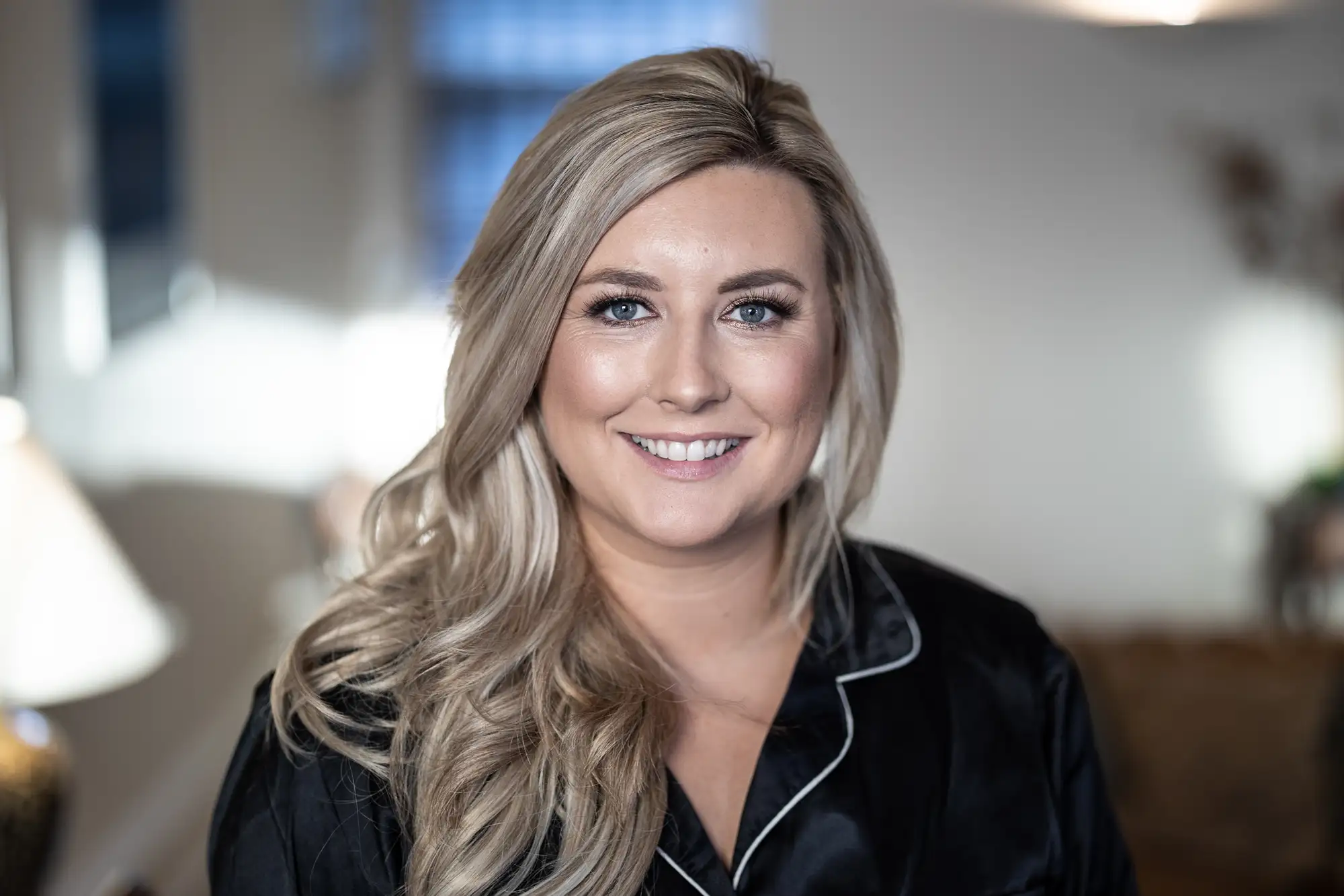 A smiling woman with blonde wavy hair, wearing a black satin shirt, indoors with soft focus background featuring home decor.