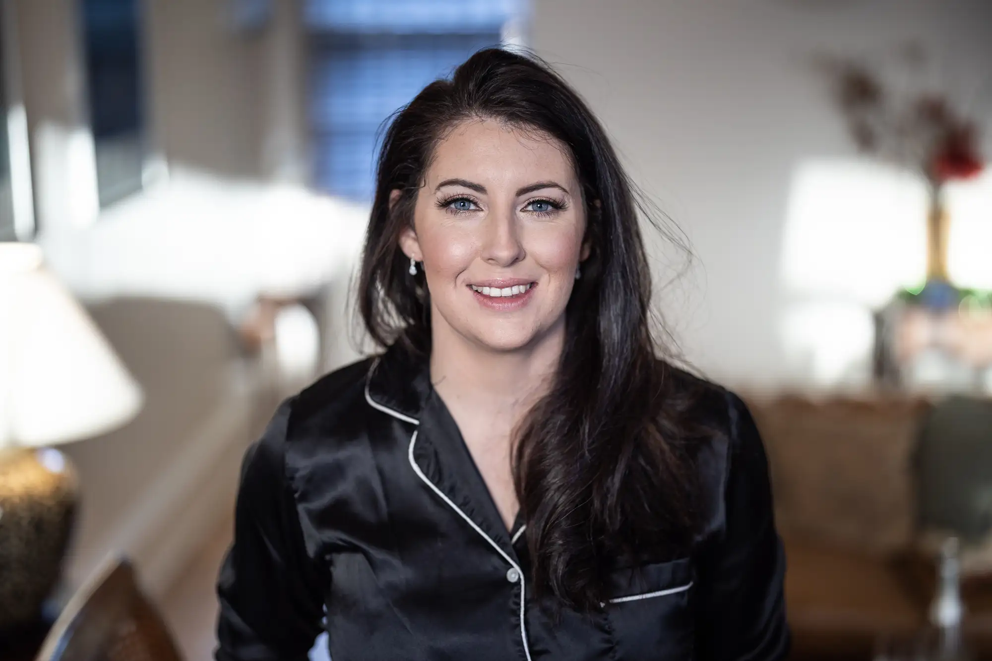 A woman with dark hair wearing a black satin pajama top smiles while seated indoors, with a softly blurred living room background.