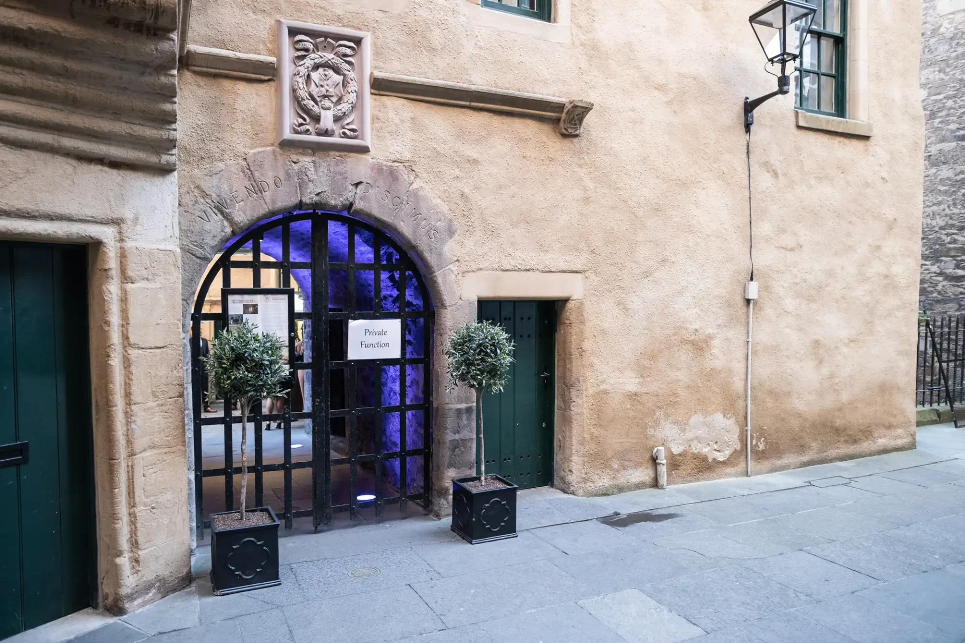 An arched entrance with a blue-lit interior, flanked by potted plants, in a weathered beige building with a crest and green doors. a sign reads "private function.