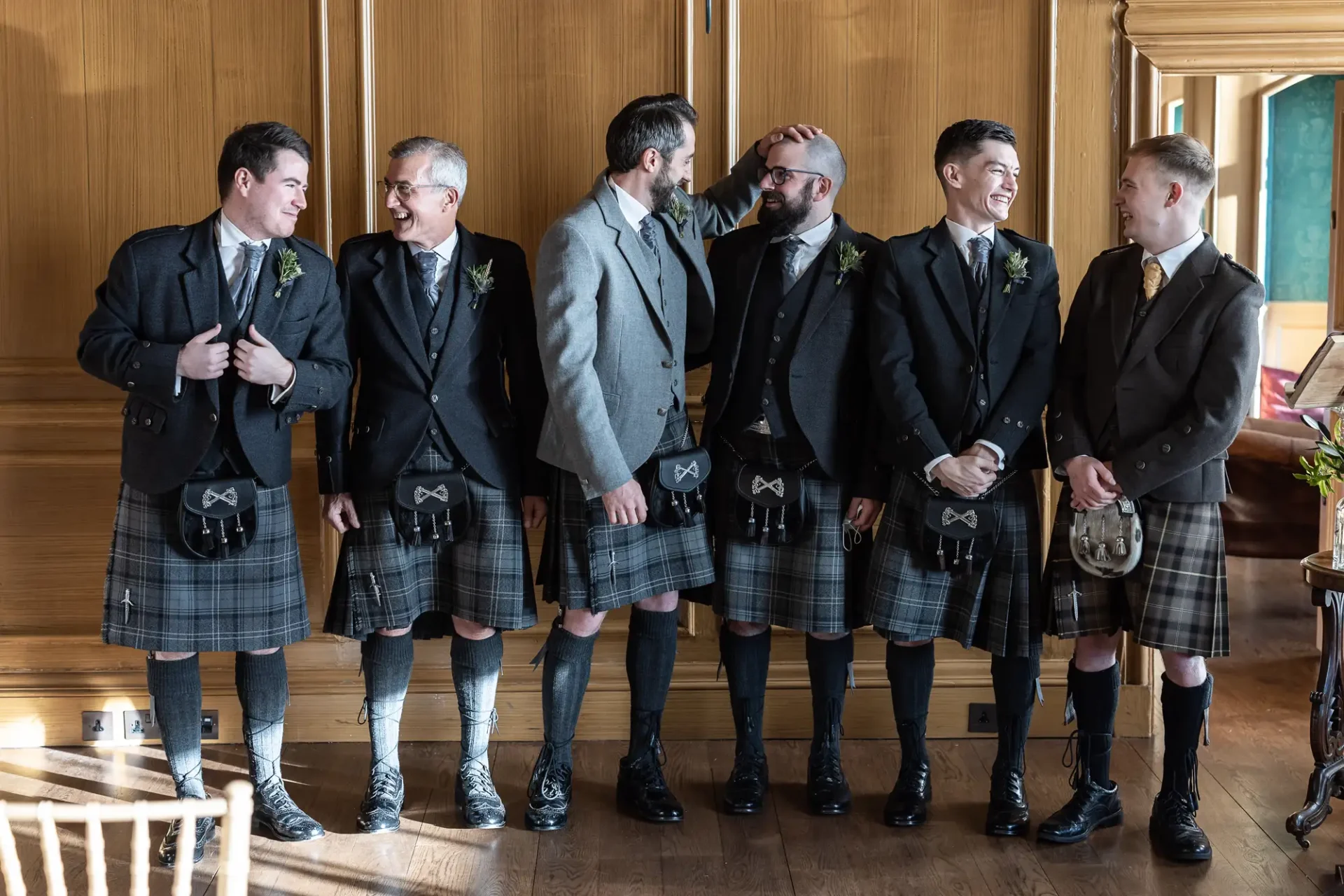 Six men in traditional scottish kilts and jackets, smiling and interacting jovially in a well-lit wooden interior.
