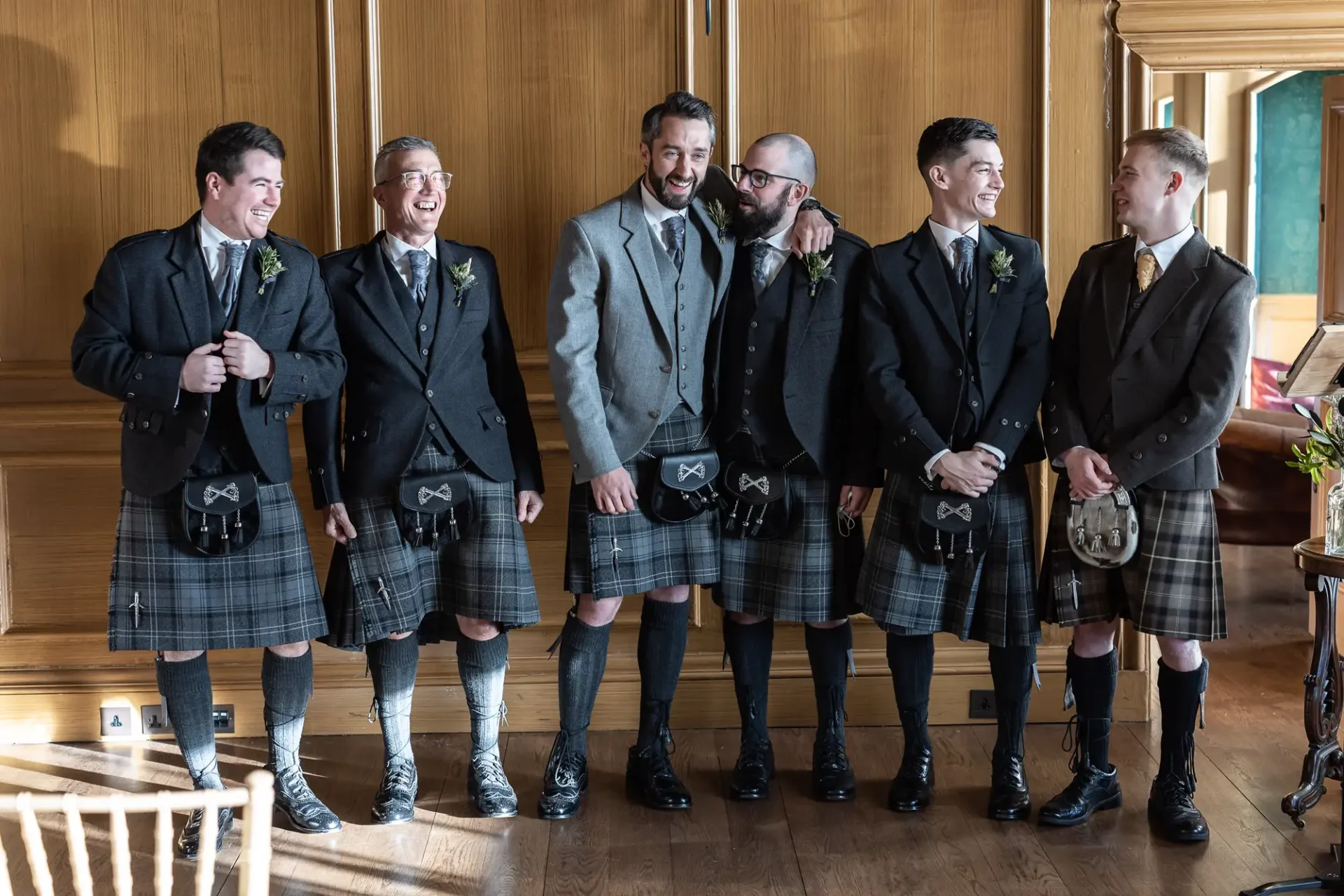 Six men in traditional tartan kilts and jackets, laughing and enjoying a moment together in a wood-paneled room.