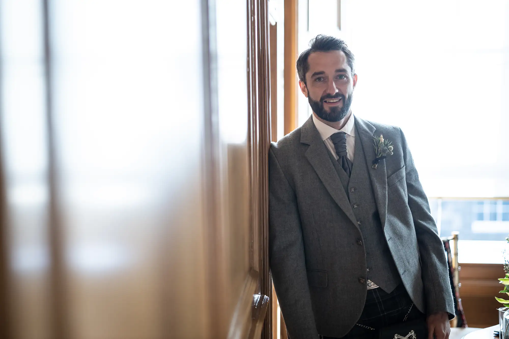 A man in a tailored gray suit and boutonniere smiling indoors, standing by a sunlit window.